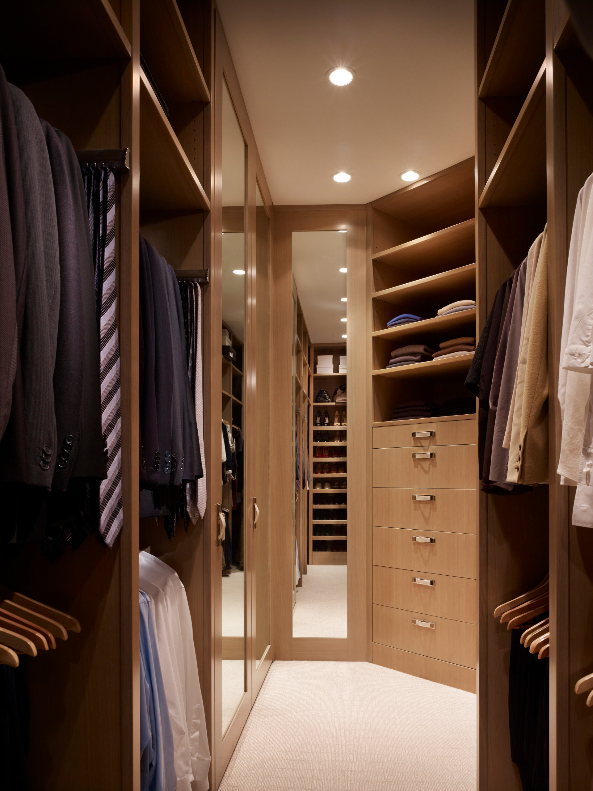 Narrow walk-in closet with warm walnut built-ins, mirrored panels, recessed lighting, and organized sections for suits, shirts, and shoes.