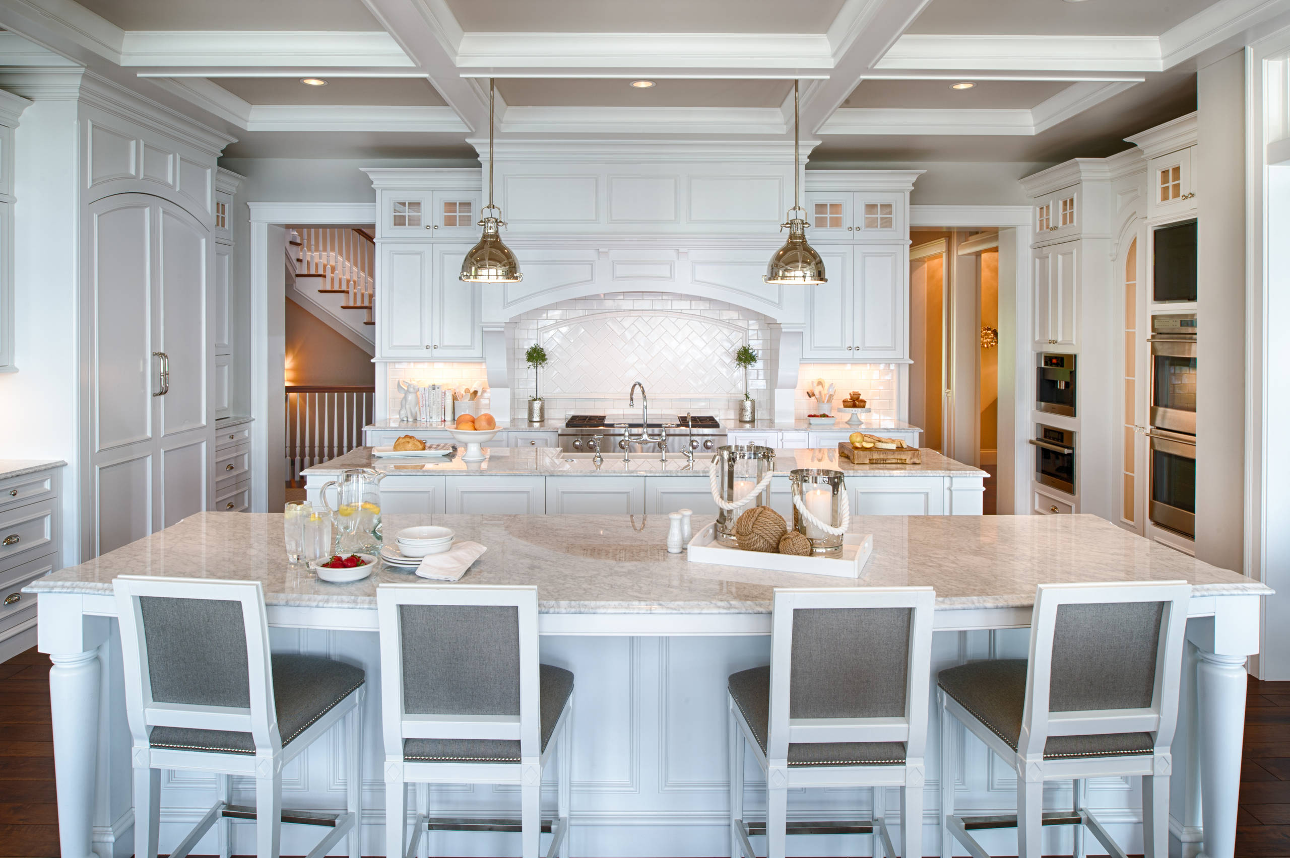 Bright white kitchen featuring expansive marble island, coffered ceiling, industrial pendants, and herringbone backsplash with gray upholstered barstools.