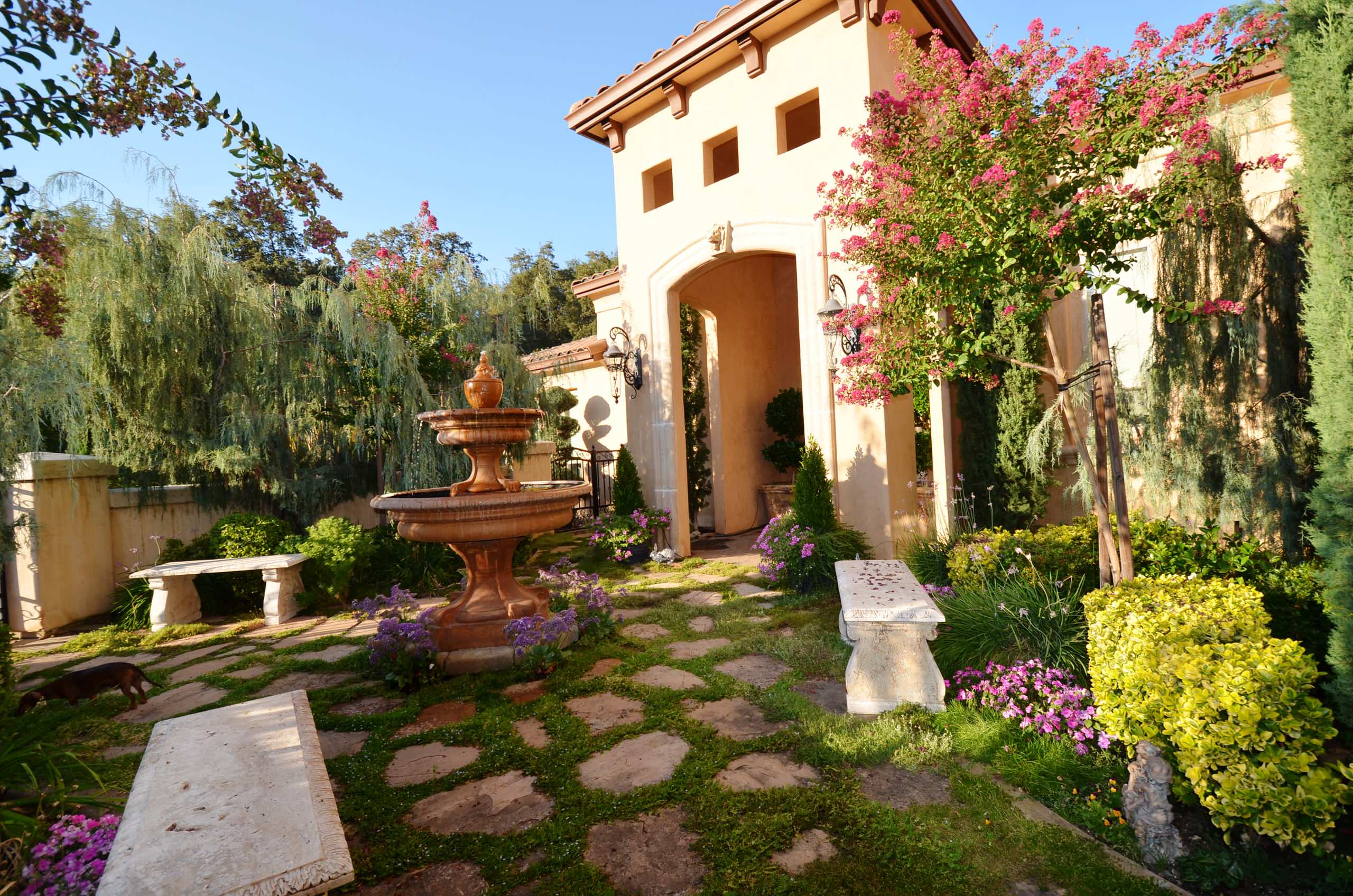 Mediterranean courtyard with tiered fountain, stone pathway, stone benches, and climbing bougainvillea against cream stucco walls.