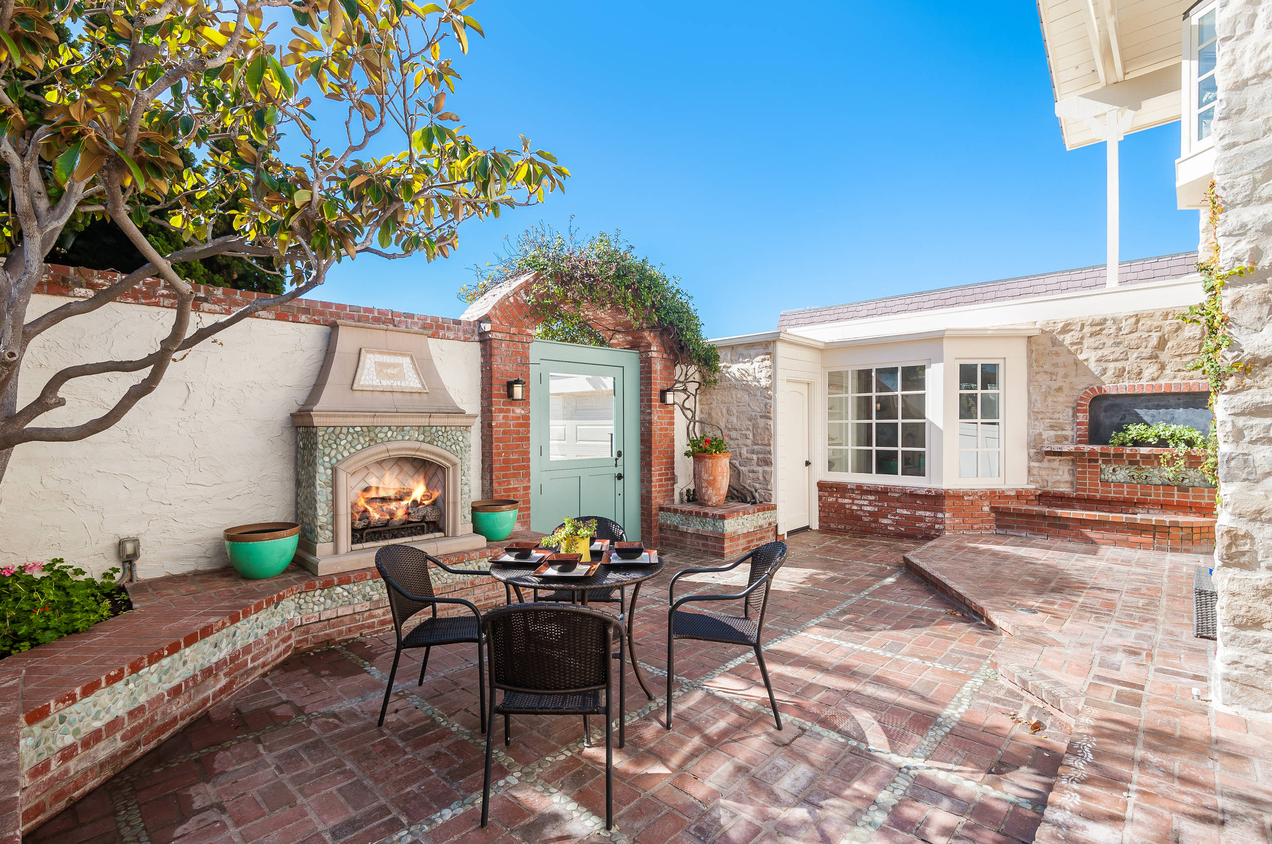 Brick-paved courtyard featuring outdoor fireplace, black wicker dining set, mint green door, and climbing vines under blue sky.