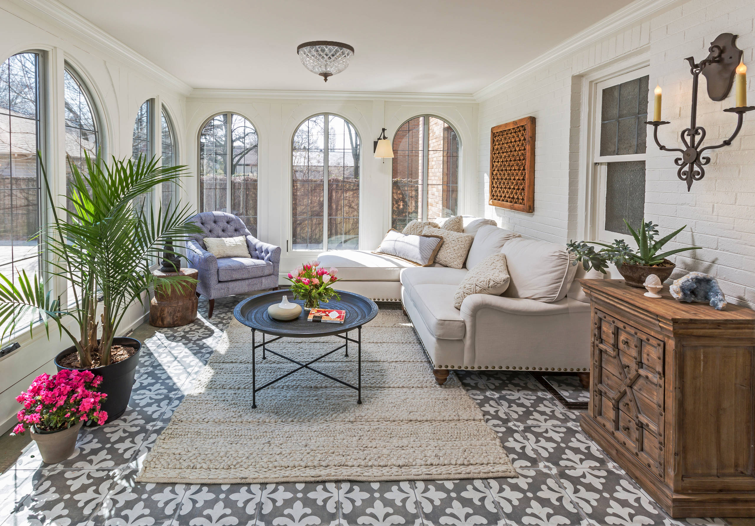 Bright sunroom with arched windows, cream sofa, blue armchair, patterned floor tiles, and tropical plants on white brick walls.
