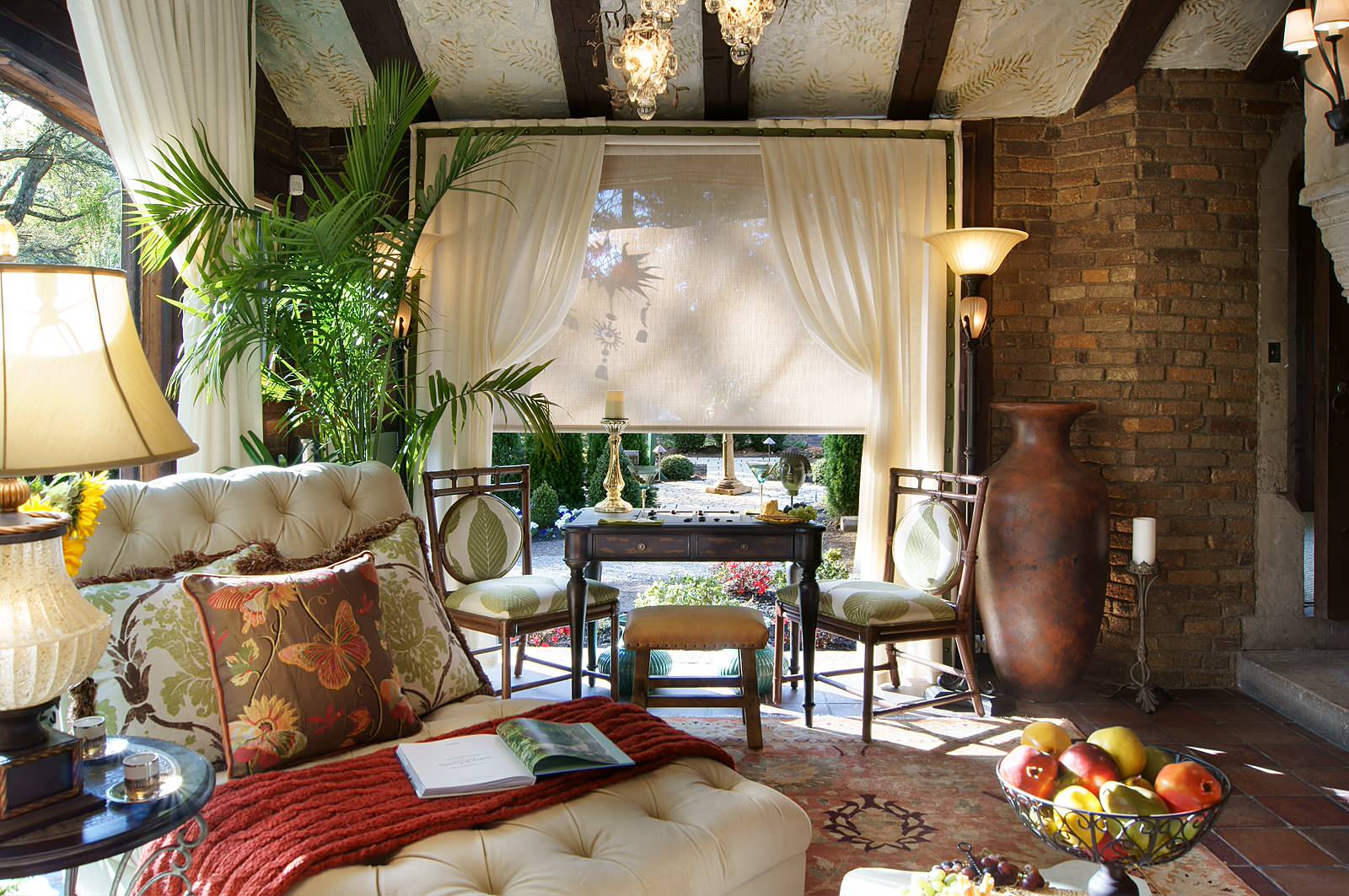 Sunlit Mediterranean sunroom featuring tufted cream chaise, palm plants, brick walls, and crystal chandeliers with flowing white curtains.