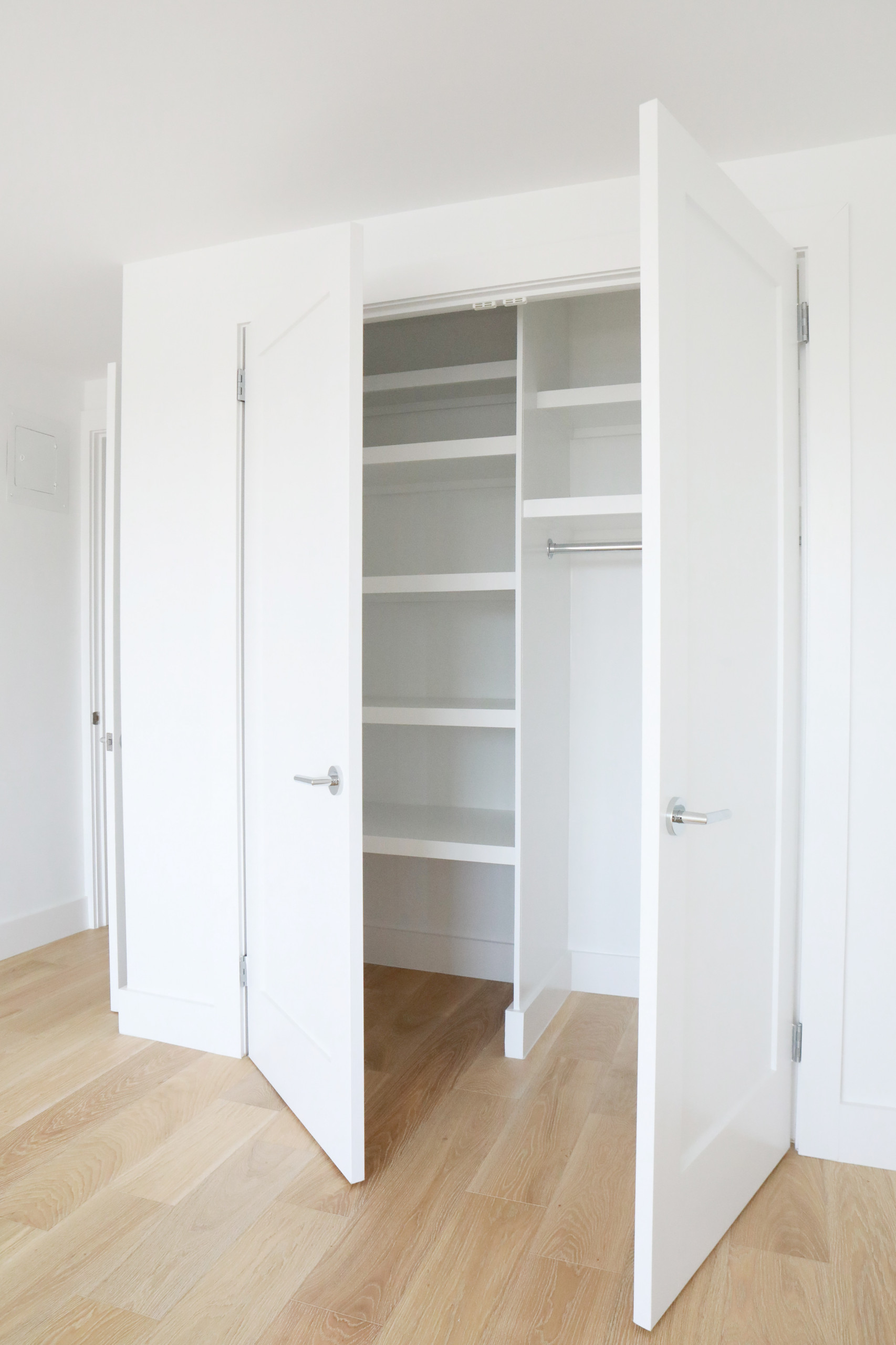 Built-in closet with matte white doors open to reveal interior shelving and hanging rod, set against light oak hardwood flooring.