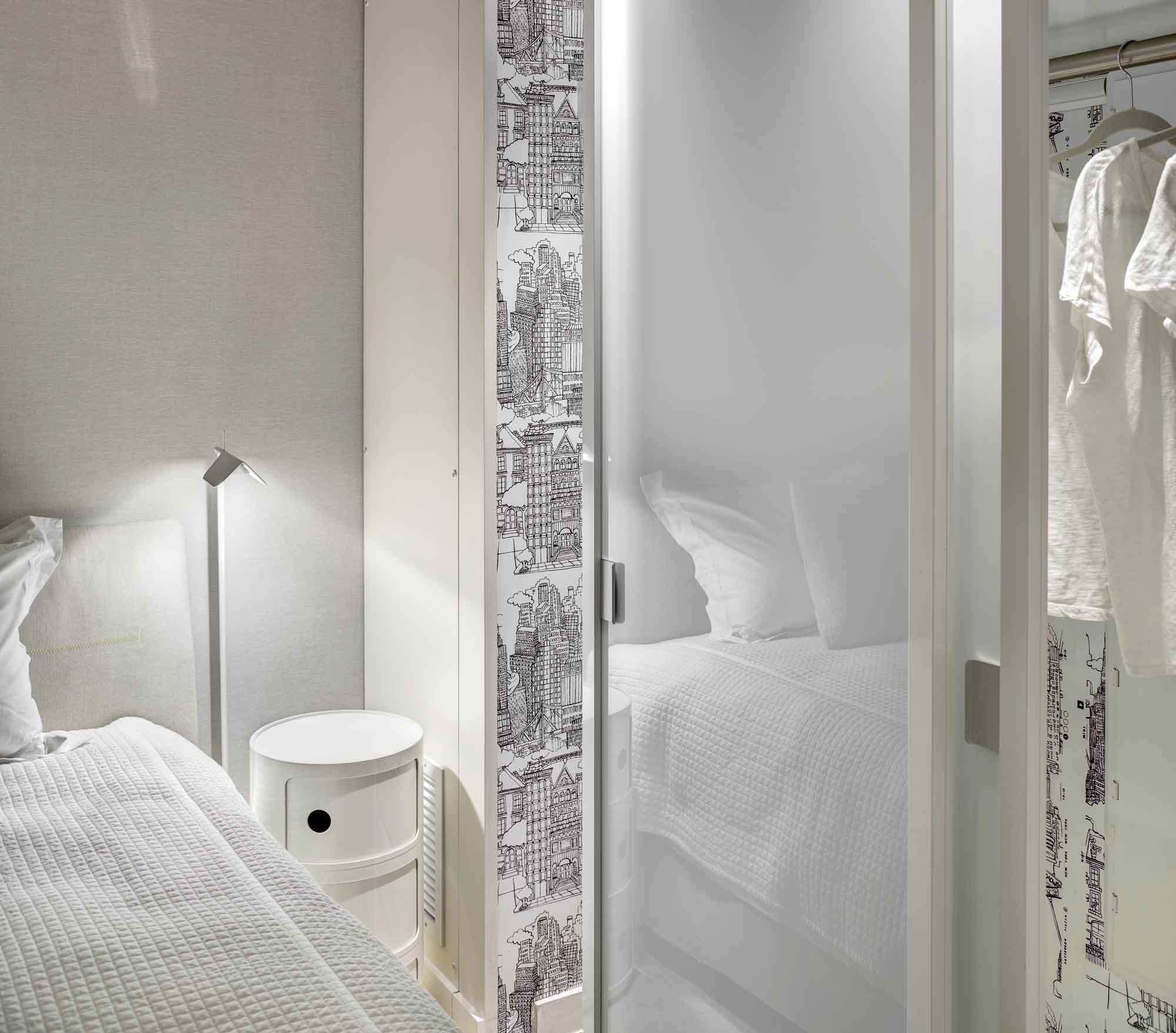 White bedroom with textured bedding, cylindrical nightstand, modern lamp, and sliding door featuring architectural sketch pattern.