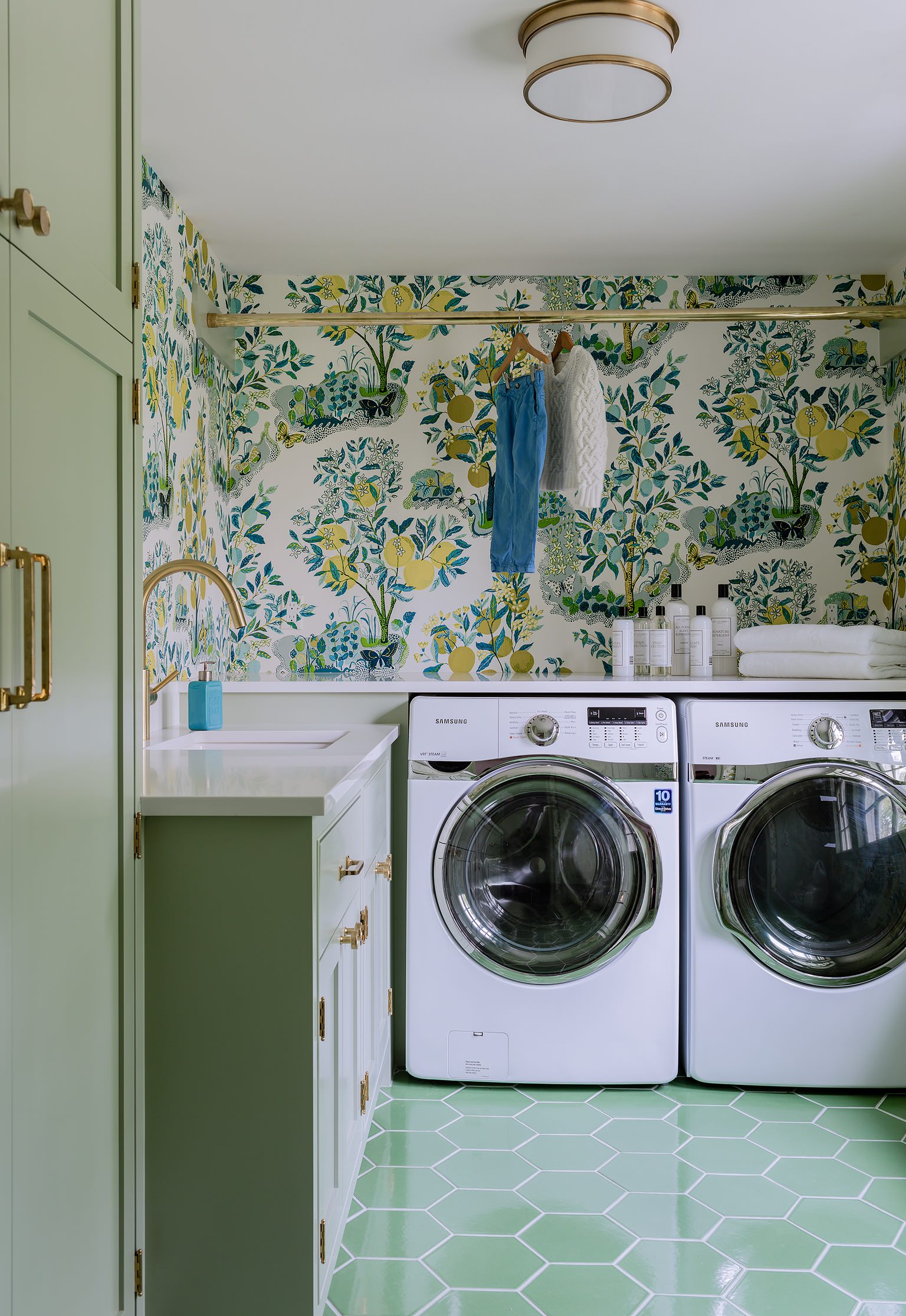 Laundry room with citrus-patterned wallpaper, mint green hexagonal floor tiles, white cabinets, and brass fixtures.