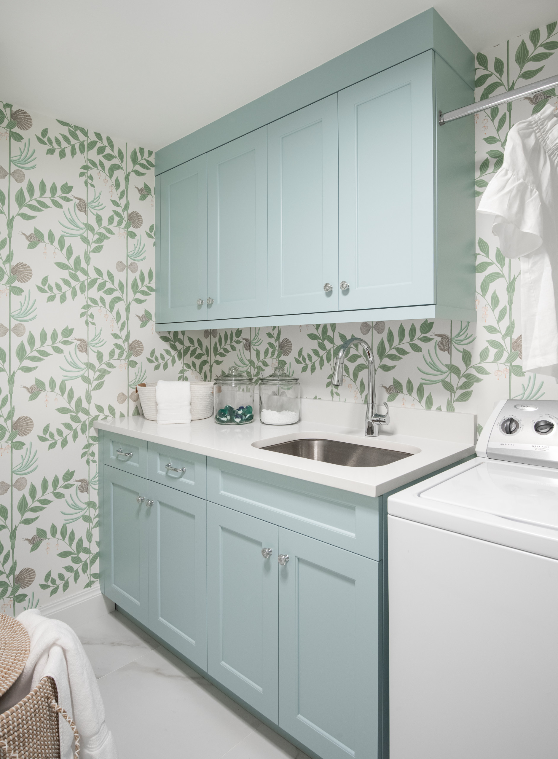 Laundry room with mint green shaker cabinets, botanical print wallpaper, white countertops, and stainless steel sink.