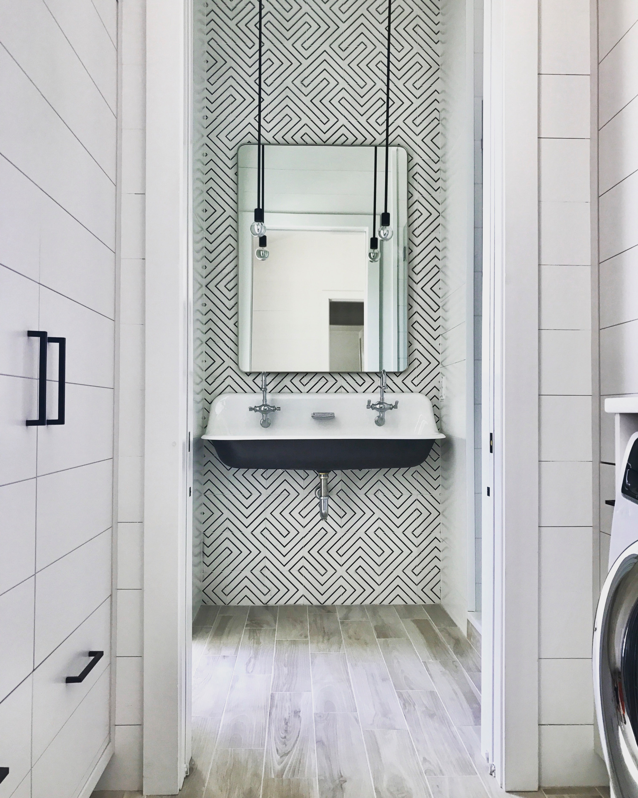 Modern bathroom featuring black and white geometric wallpaper, wall-mounted trough sink, pendant lights, and light wood-look flooring.
