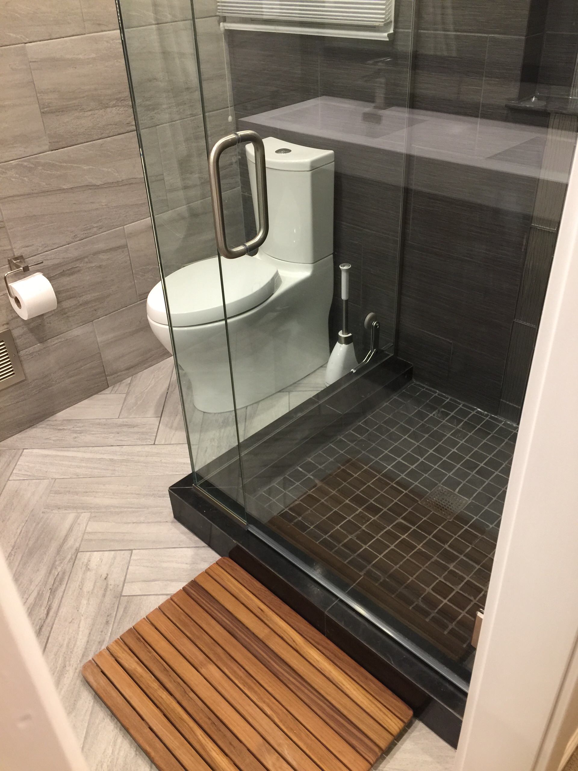 Modern bathroom featuring glass shower enclosure, teak floor mat, gray herringbone tile, and black mosaic shower floor.