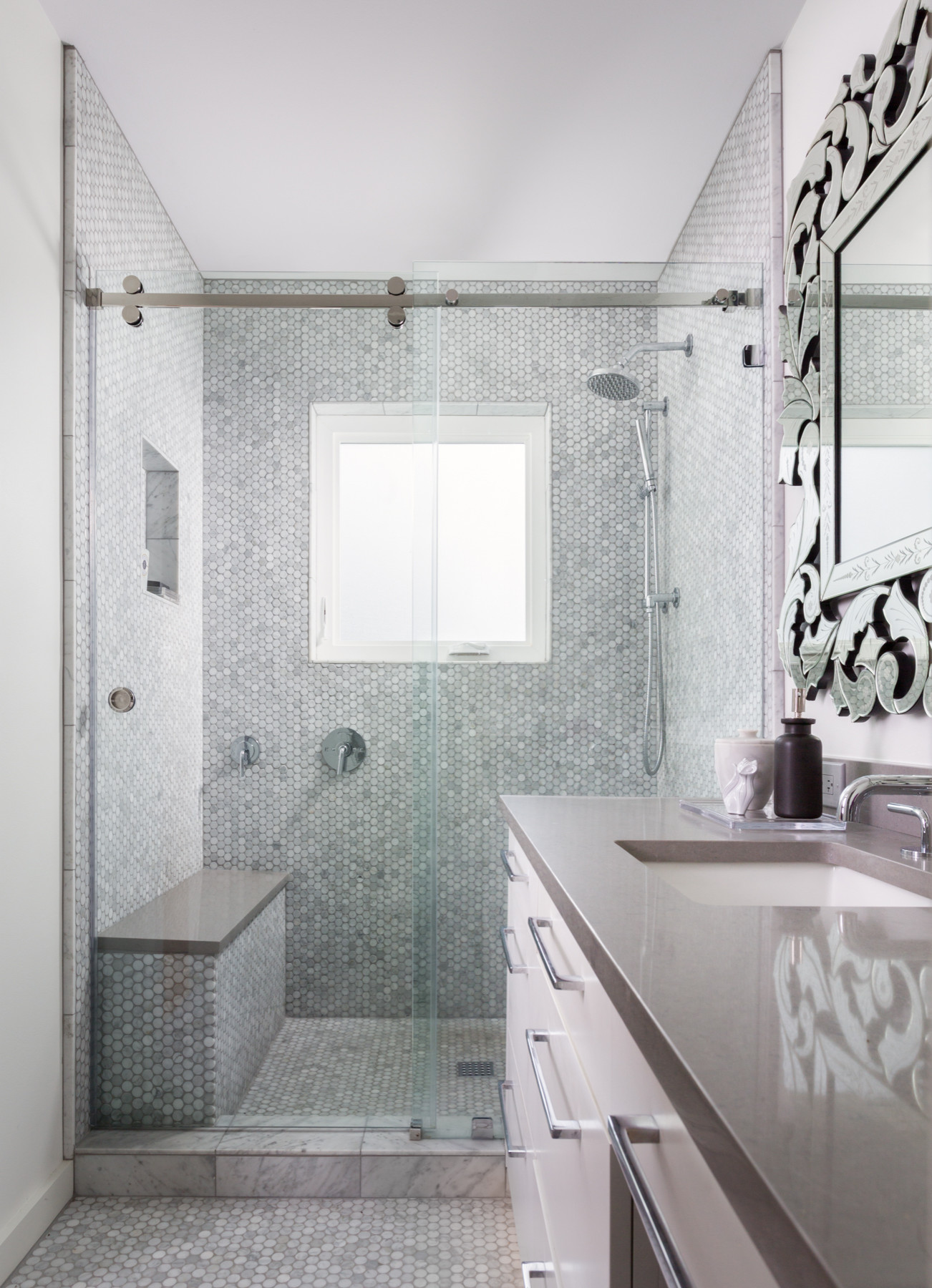 Modern bathroom featuring glass-enclosed shower with hexagonal mosaic tiles, floating vanity, and ornate mirror against white walls.