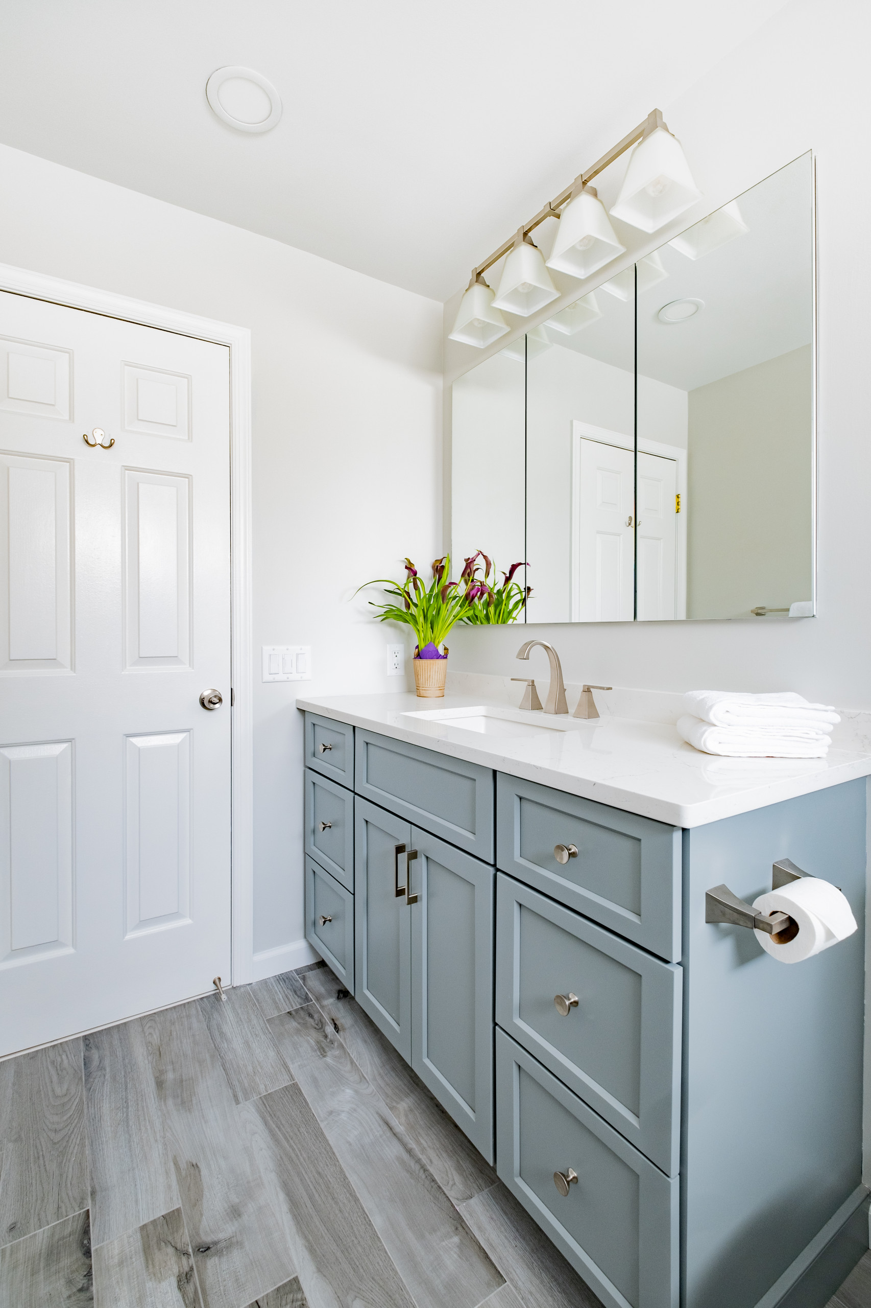 Bathroom featuring sage green vanity cabinet, white quartz countertop, triple mirror, and gray wood-look flooring with fresh flowers.