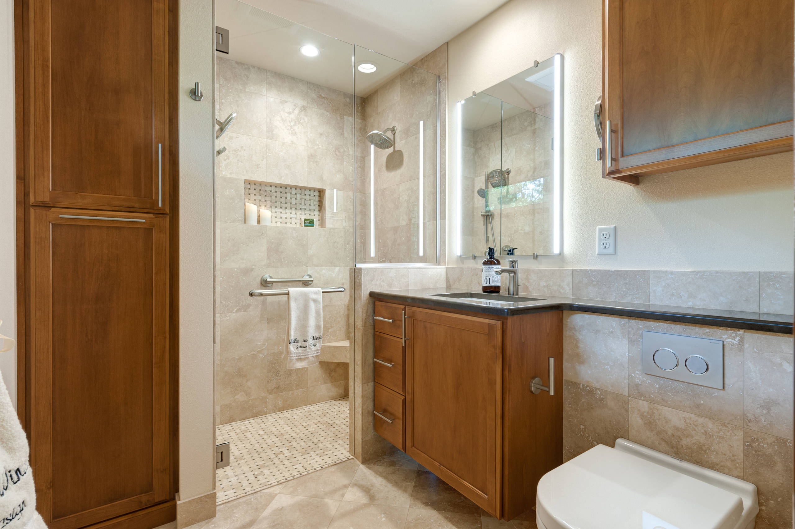 Modern bathroom featuring wooden cabinetry, beige limestone tiles, frameless glass shower, and LED-lit mirrors with black countertops.
