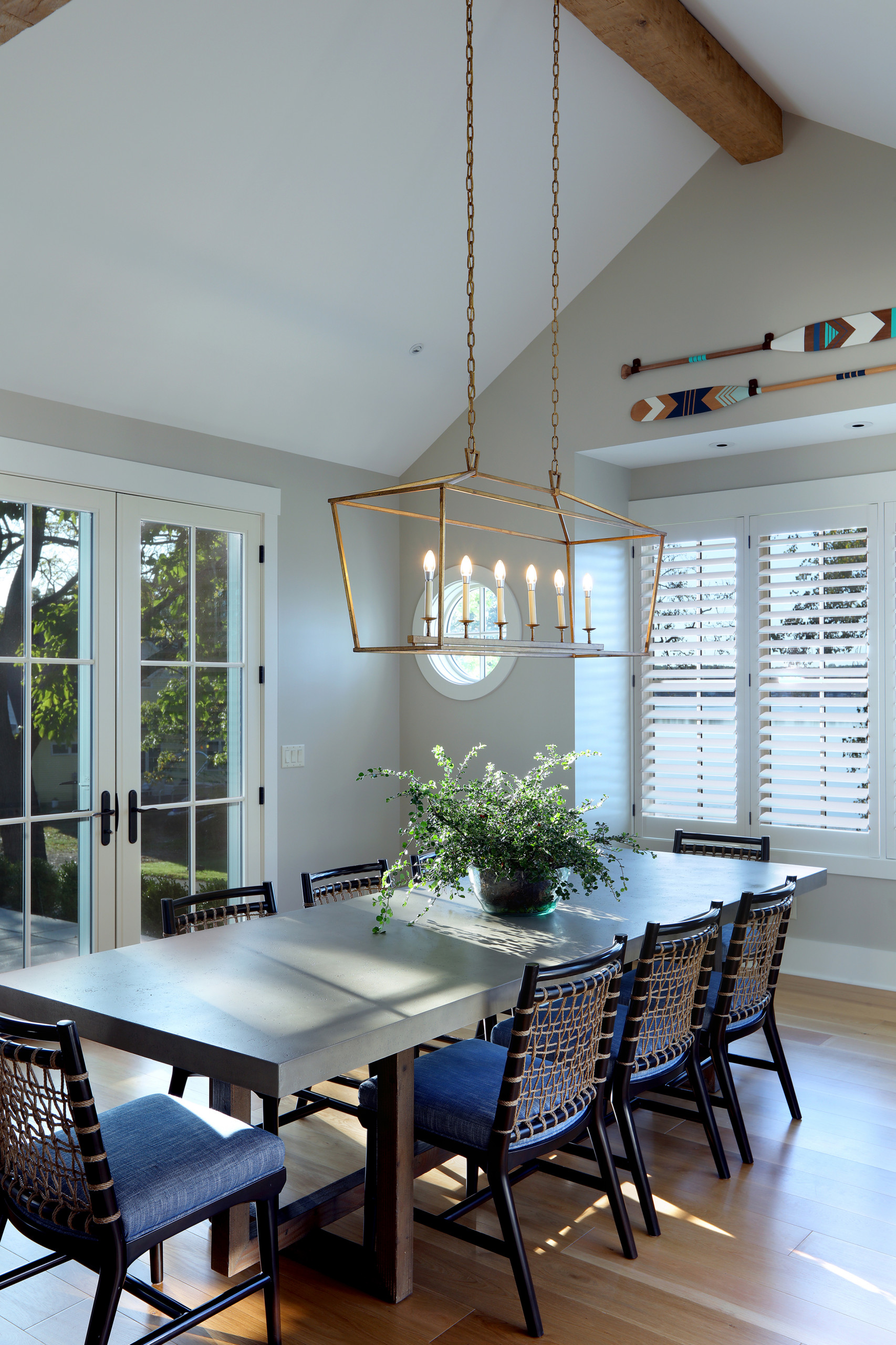 Bright dining room with concrete table, woven chairs, brass linear chandelier, French doors, and decorative oars on wall.