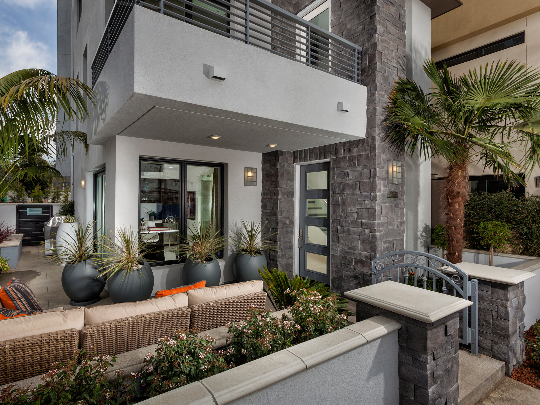 Modern home entrance featuring gray stone columns, glass doors, wicker seating, and spherical planters with tropical landscaping.