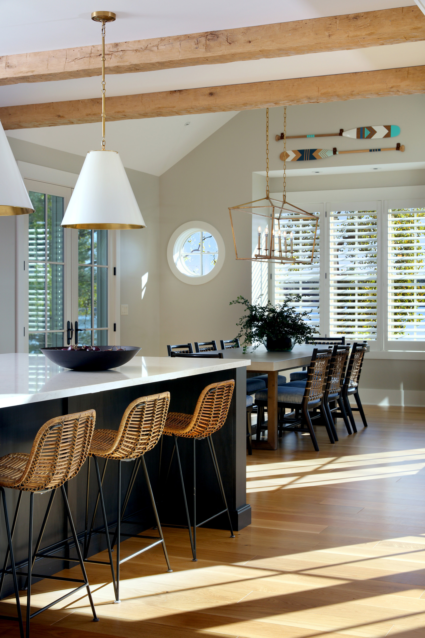 Open kitchen with rattan barstools, exposed wooden beams, white pendant lights, and plantation shutters filtering natural light.