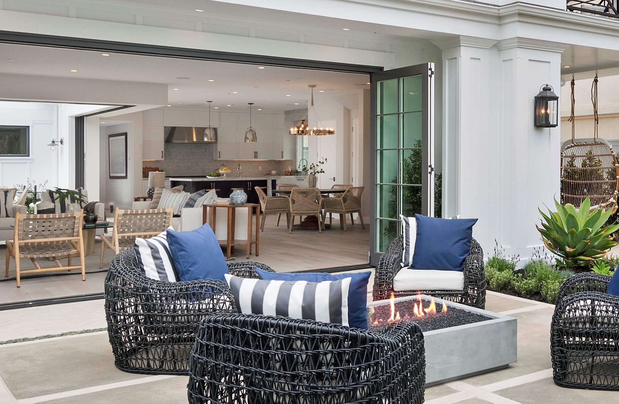 Modern patio with woven chairs, blue cushions, concrete fire pit, and folding glass doors leading to bright kitchen interior.