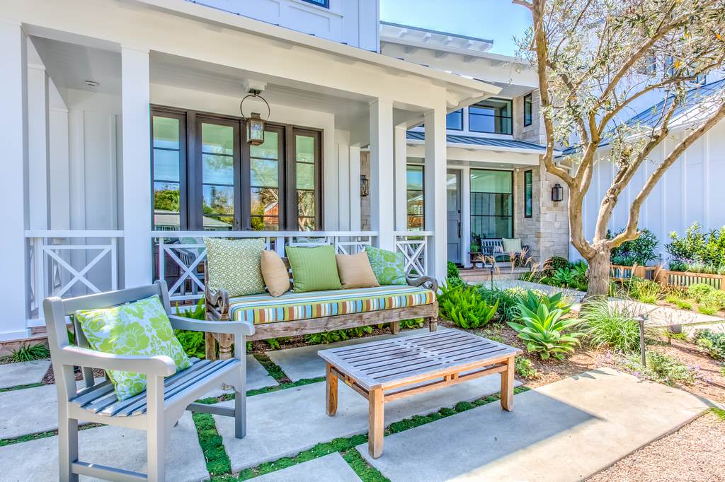 White coastal porch with gray wooden benches, striped cushions, green pillows, and lush garden landscaping beneath mature tree.