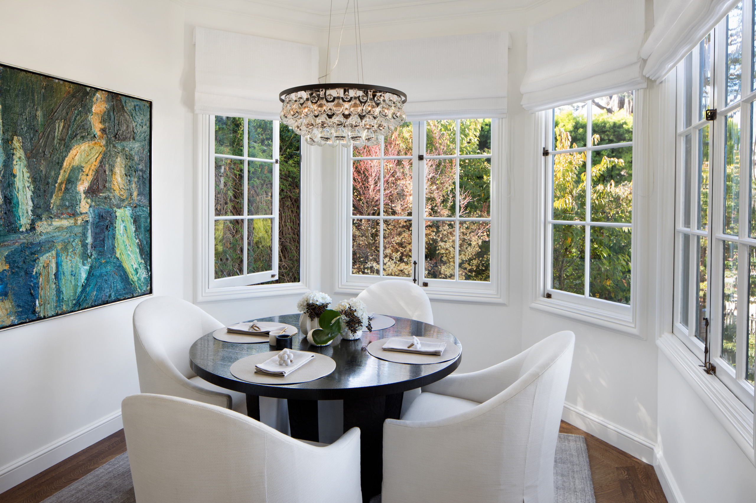 Bright dining nook with round black table, white upholstered chairs, crystal chandelier, and wraparound windows overlooking garden.