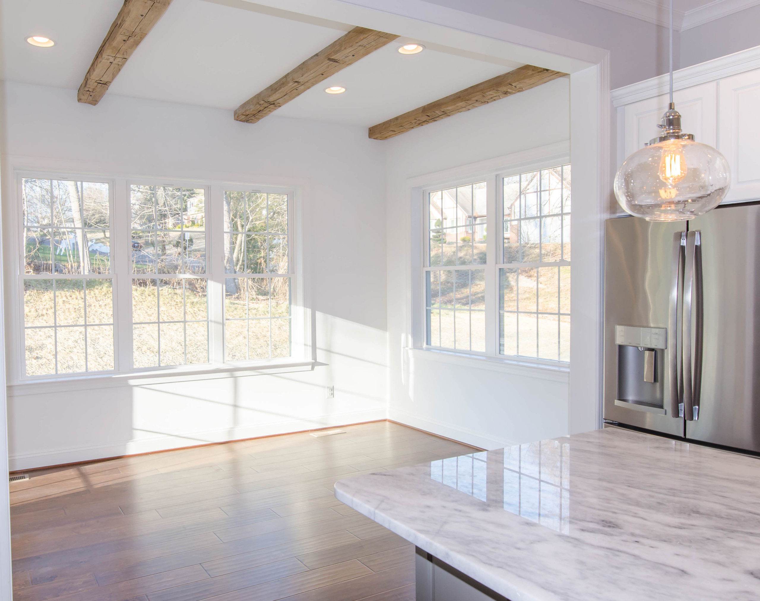 Bright kitchen space with exposed wooden beams, large windows, marble countertop, and hardwood floors bathed in natural light.