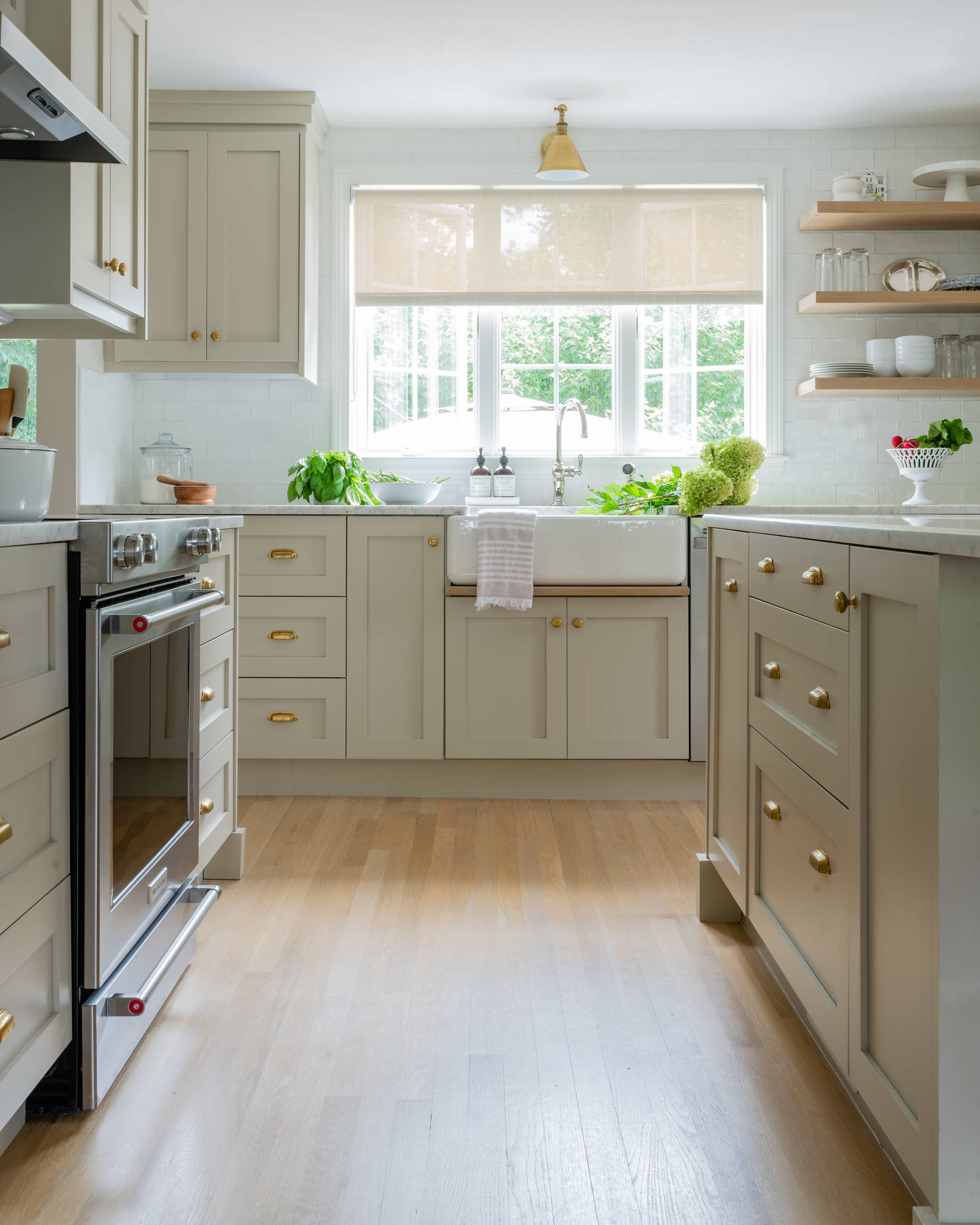 Sage green kitchen with farmhouse sink, floating wood shelves, large window, and brass hardware against white walls.