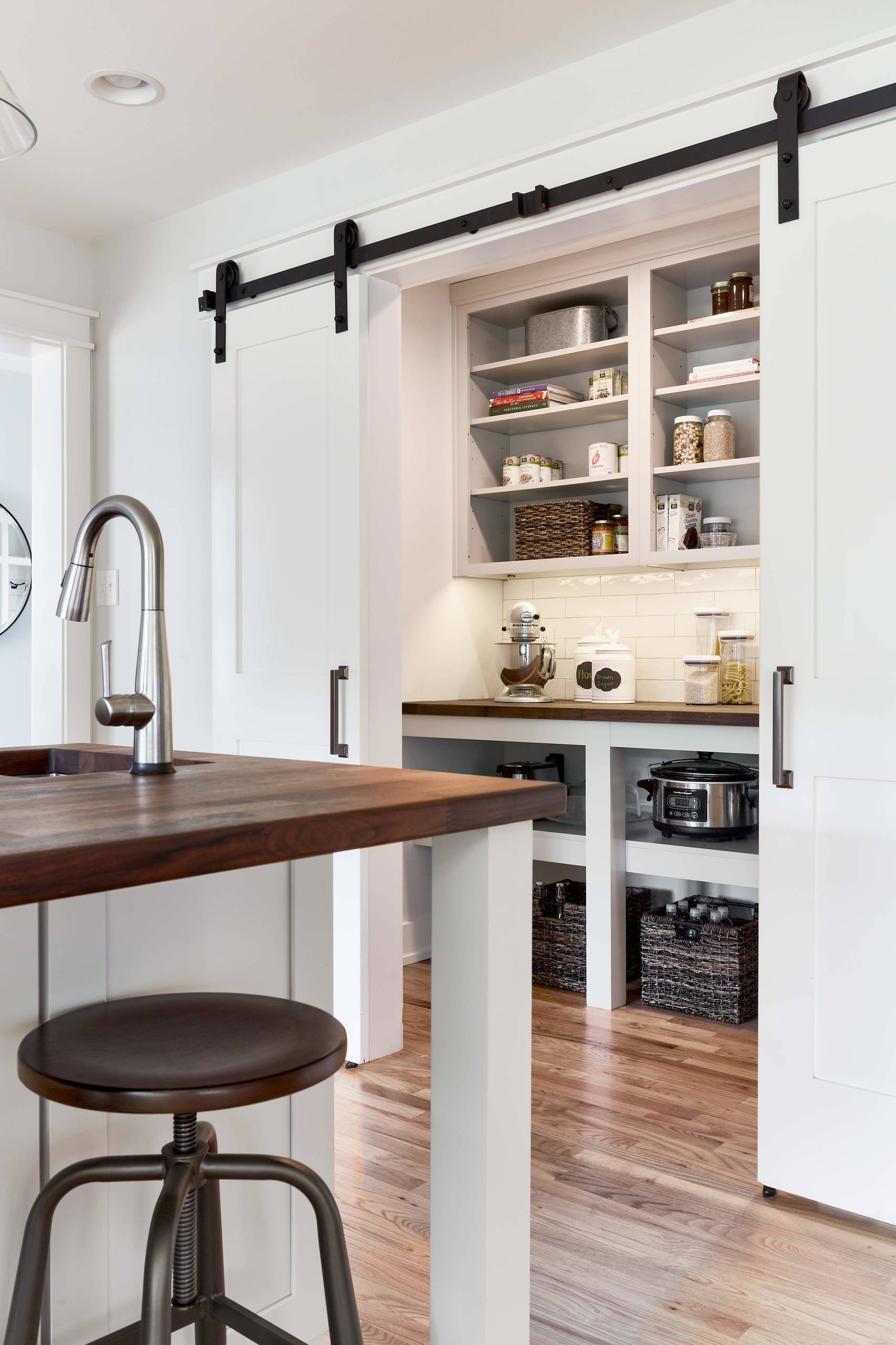 Modern kitchen pantry with black barn door hardware, white shelving, butcher block counter, and industrial stool beside stainless faucet.