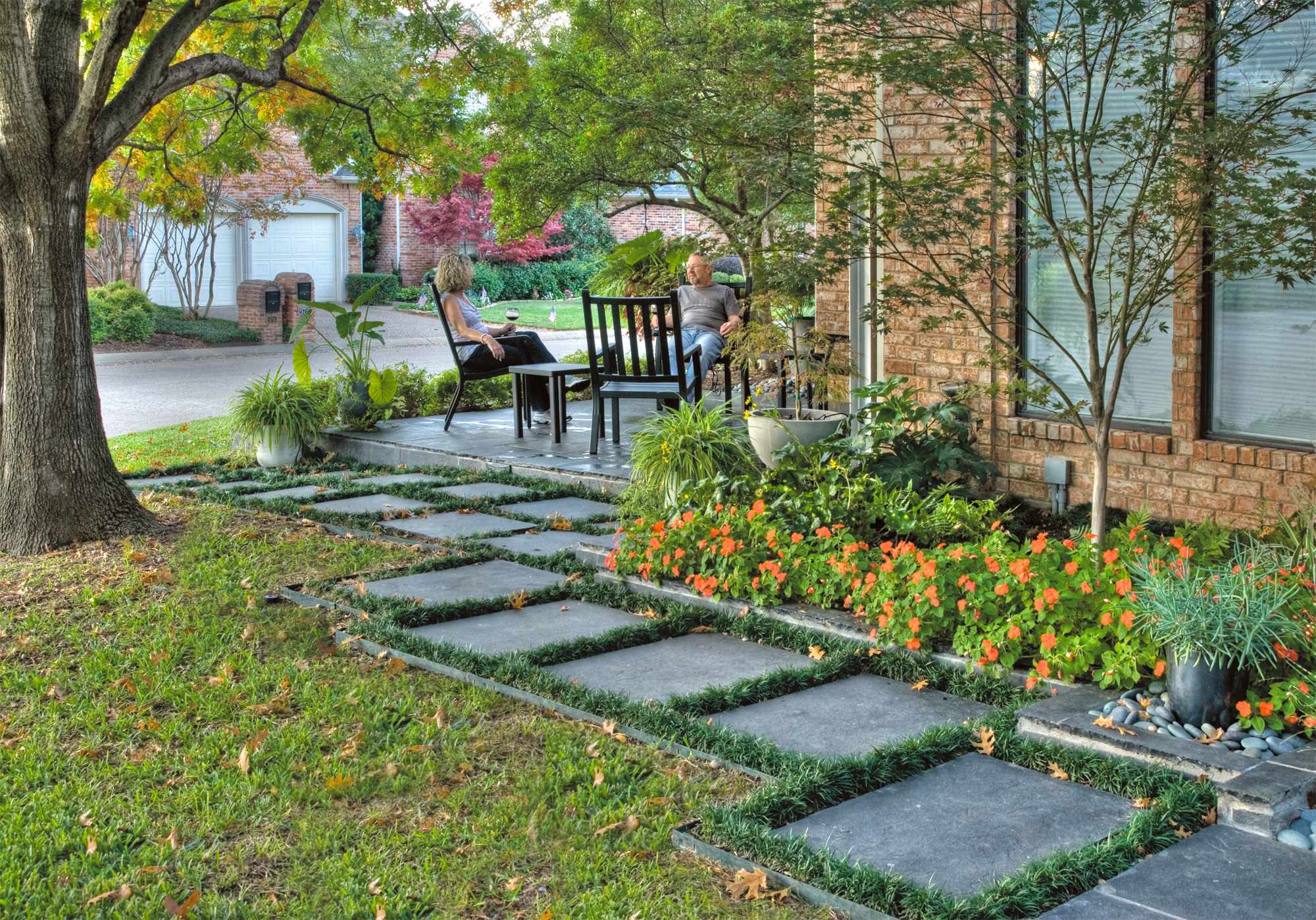 Modern stepping stone pathway with grass joints leads to concrete patio, bordered by orange flowers and mature trees against brick house.