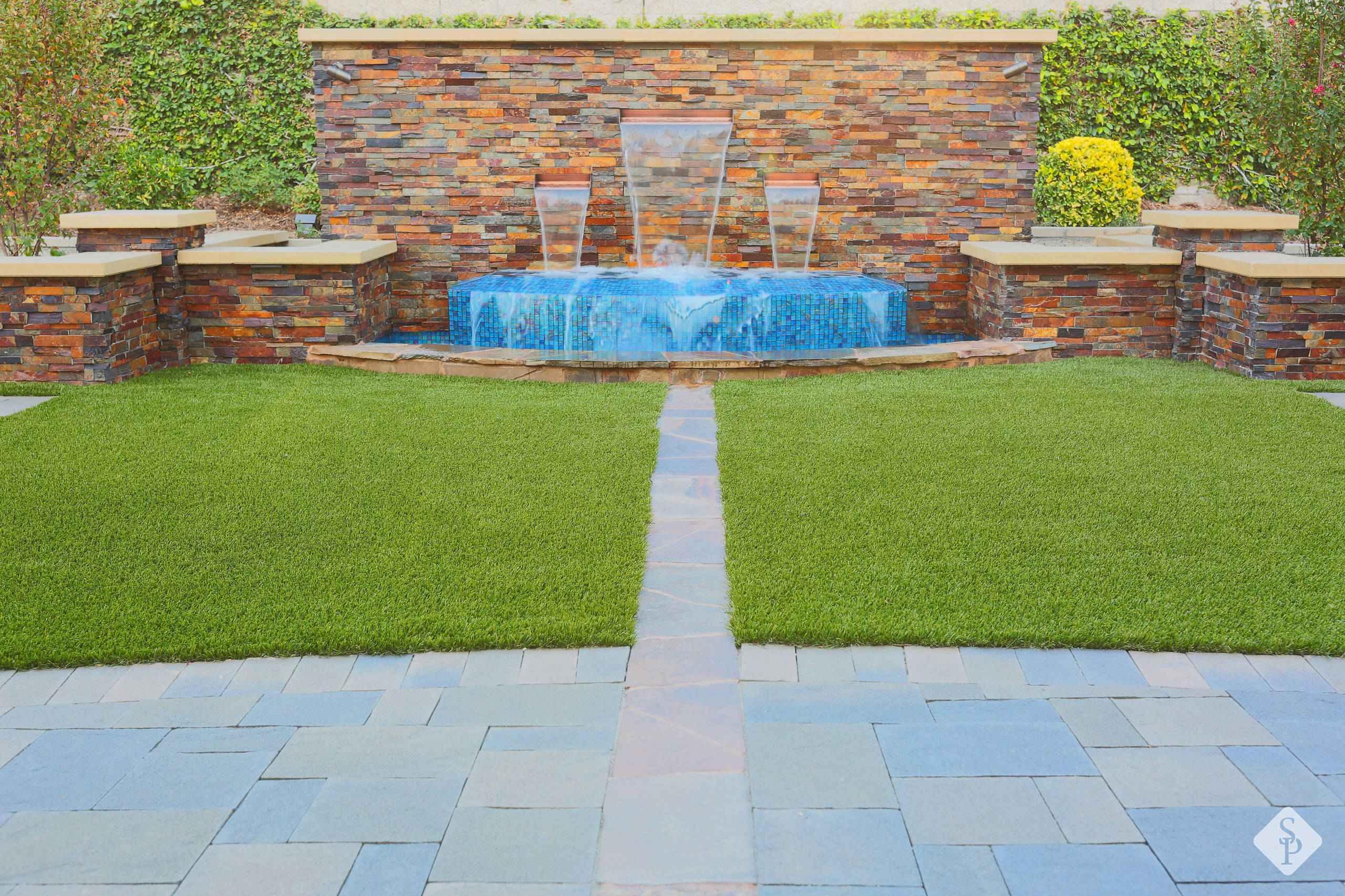 Modern garden featuring stacked stone wall with glass waterfall panels, blue mosaic tile basin, and manicured lawn with paver pathway.