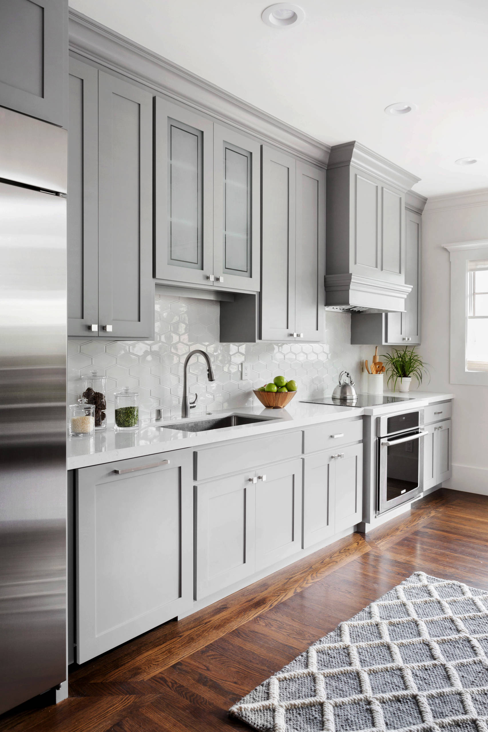 Modern kitchen with gray shaker cabinets, white hexagonal backsplash, quartz countertops, and dark hardwood floors.
