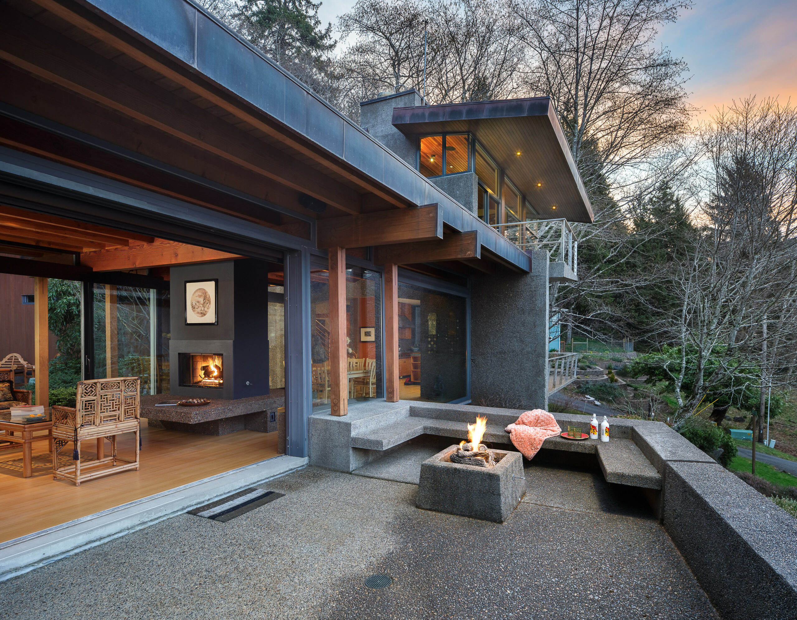 Modern terrace with concrete fire pit, built-in seating, indoor fireplace, and sliding glass walls overlooking wooded hillside at dusk.