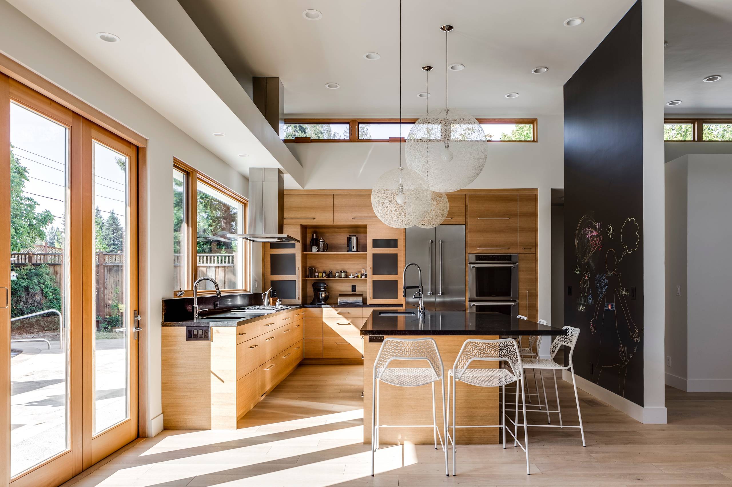Modern kitchen with light wood cabinets, black countertops, floor-to-ceiling windows, and spherical pendant lights over island.