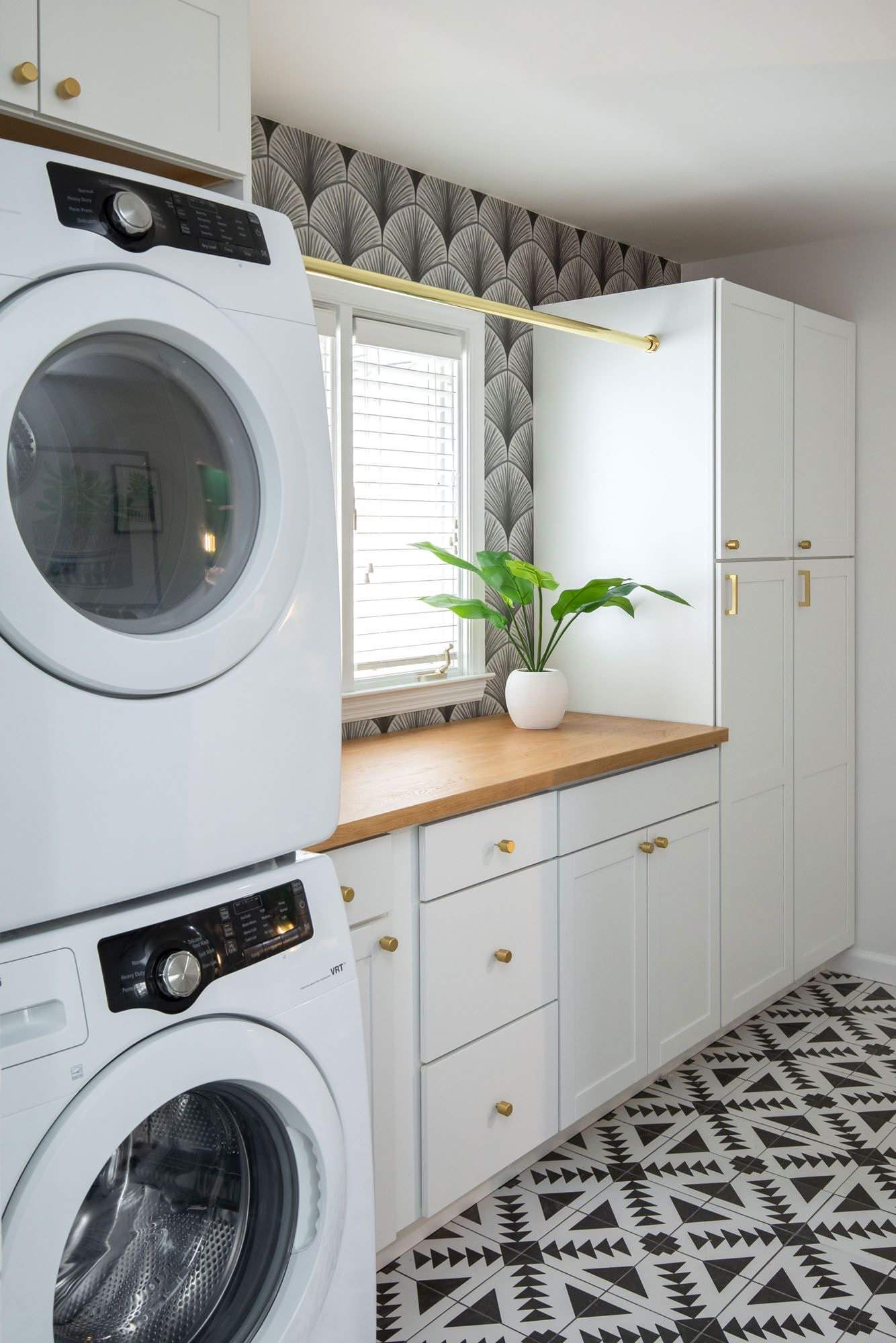 Laundry room with stacked white washers, geometric floor tile, art deco wallpaper, and white cabinets with brass hardware.