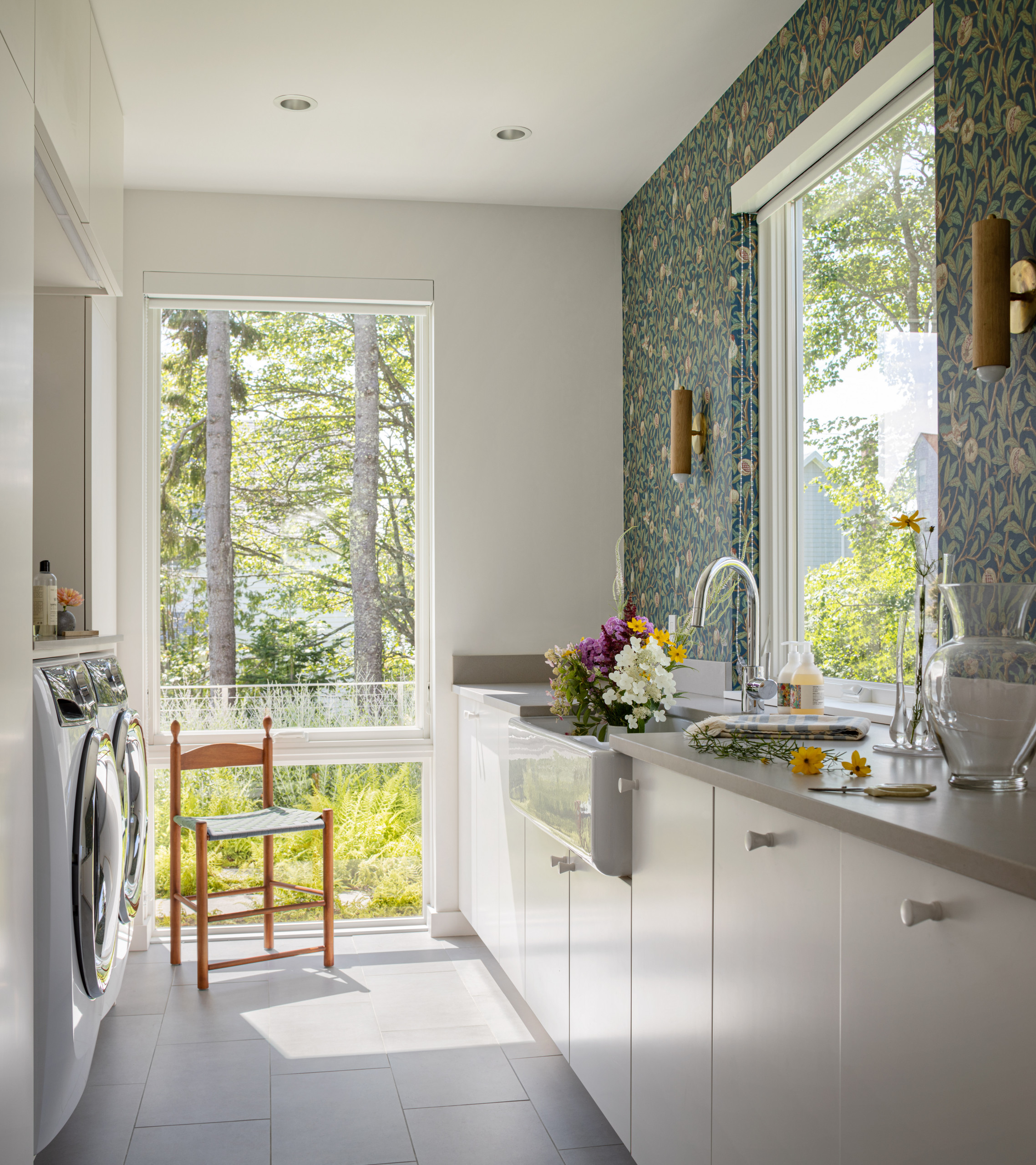 Bright laundry room with blue floral wallpaper, white cabinets, forest view windows, and wooden chair against natural scenery.