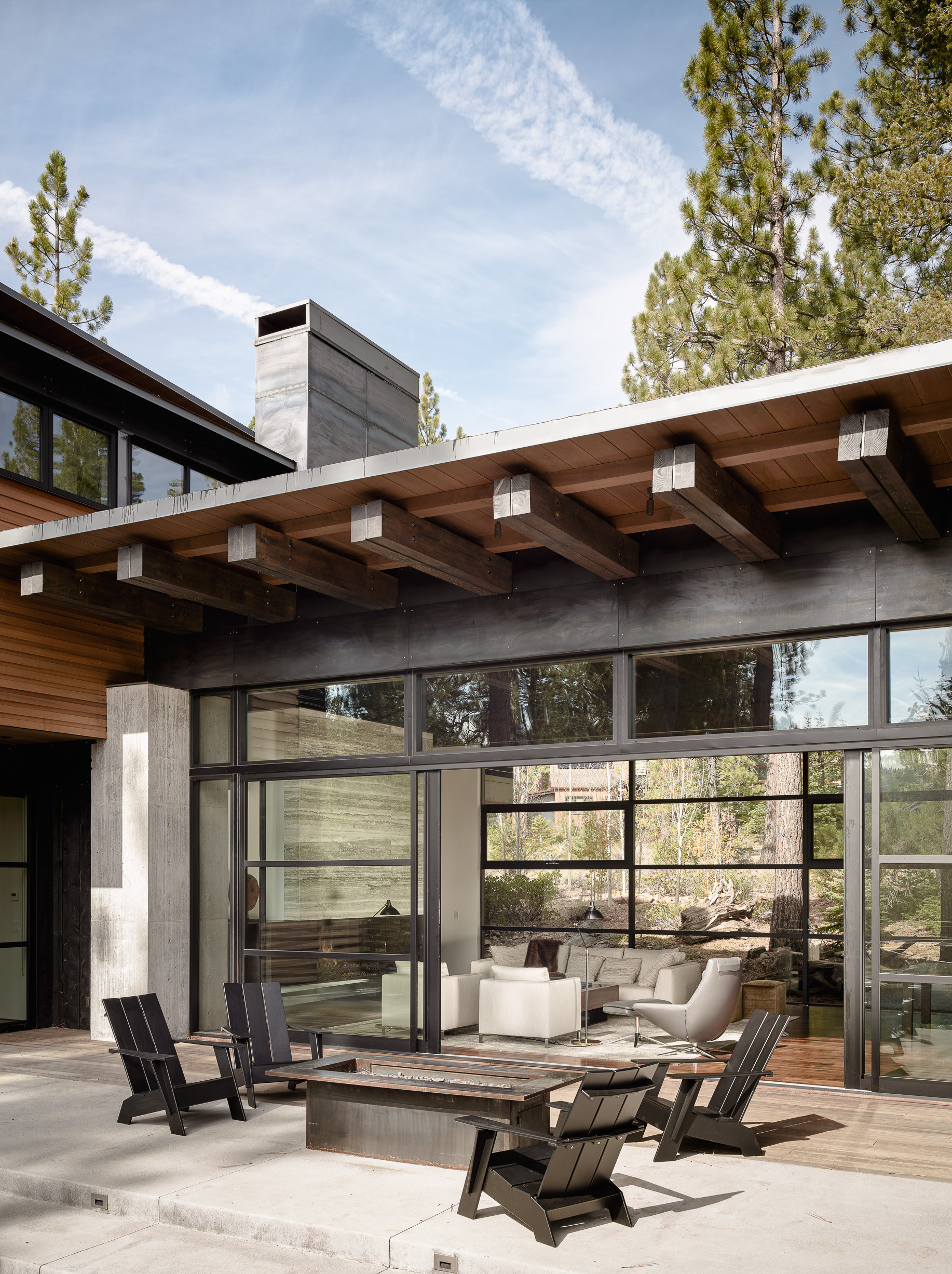 Contemporary mountain home featuring floor-to-ceiling glass walls, outdoor deck with black Adirondack chairs and linear firepit.