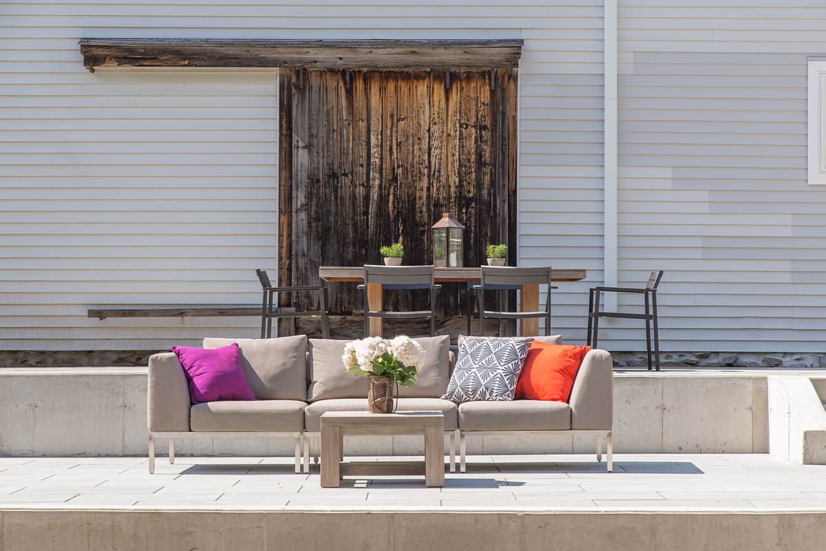 Modern outdoor seating area with gray sectional, colorful pillows, and weathered barn door backdrop against light gray siding.