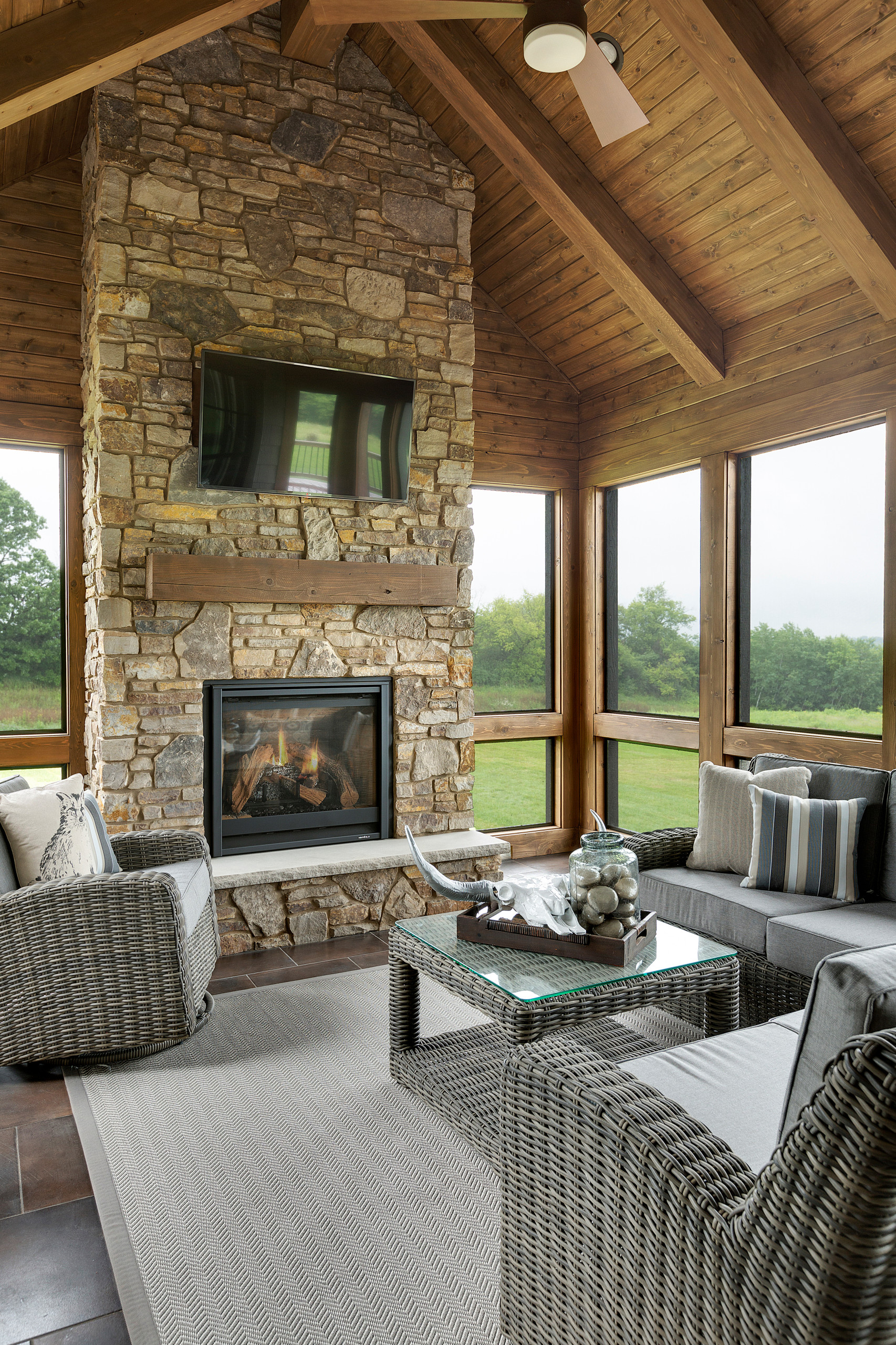 Sunroom featuring floor-to-ceiling stone fireplace, vaulted wood ceiling, panoramic windows, and gray wicker furniture set.