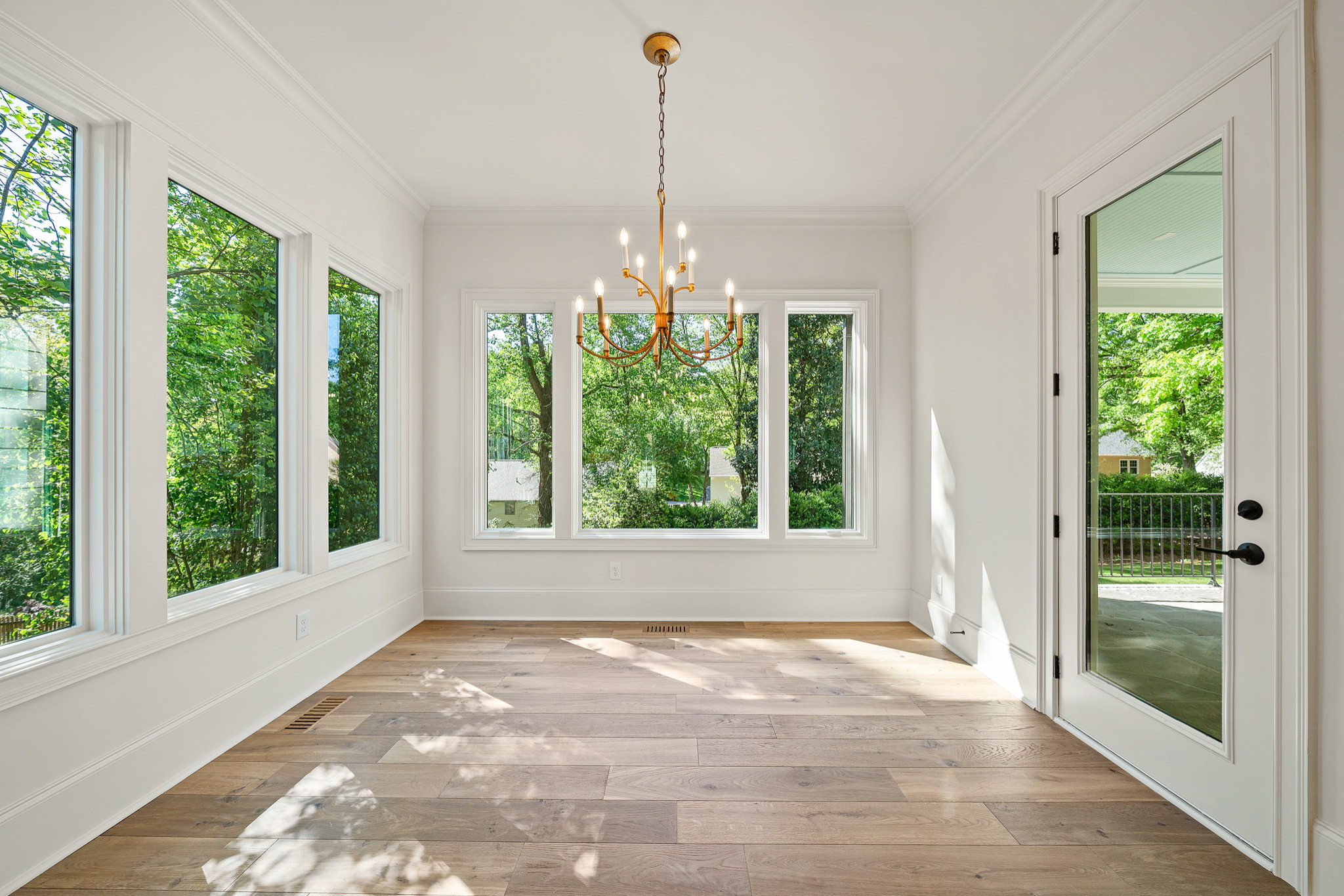 Bright sunroom with white walls, oak flooring, brass chandelier, and floor-to-ceiling windows overlooking lush greenery.