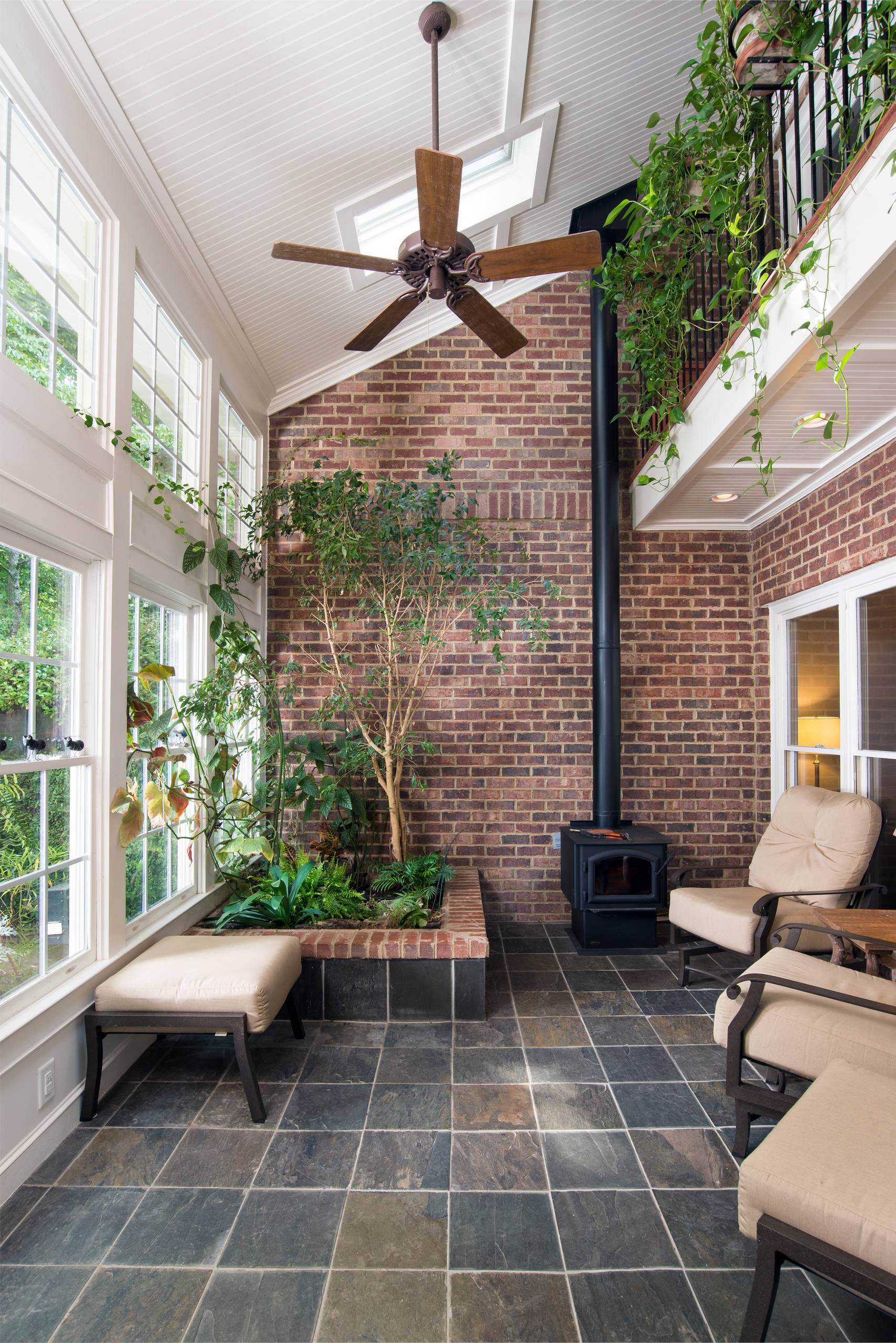 Sunroom featuring exposed brick walls, slate flooring, indoor garden bed with climbing plants, wood ceiling fan, and beige lounge chairs.