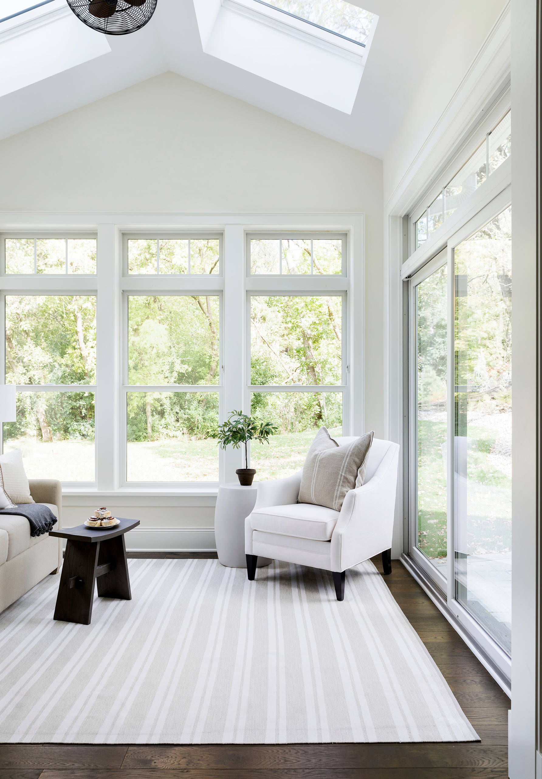 Bright sunroom featuring cathedral ceiling, floor-to-ceiling windows, white armchair, dark wood side table, and striped area rug.