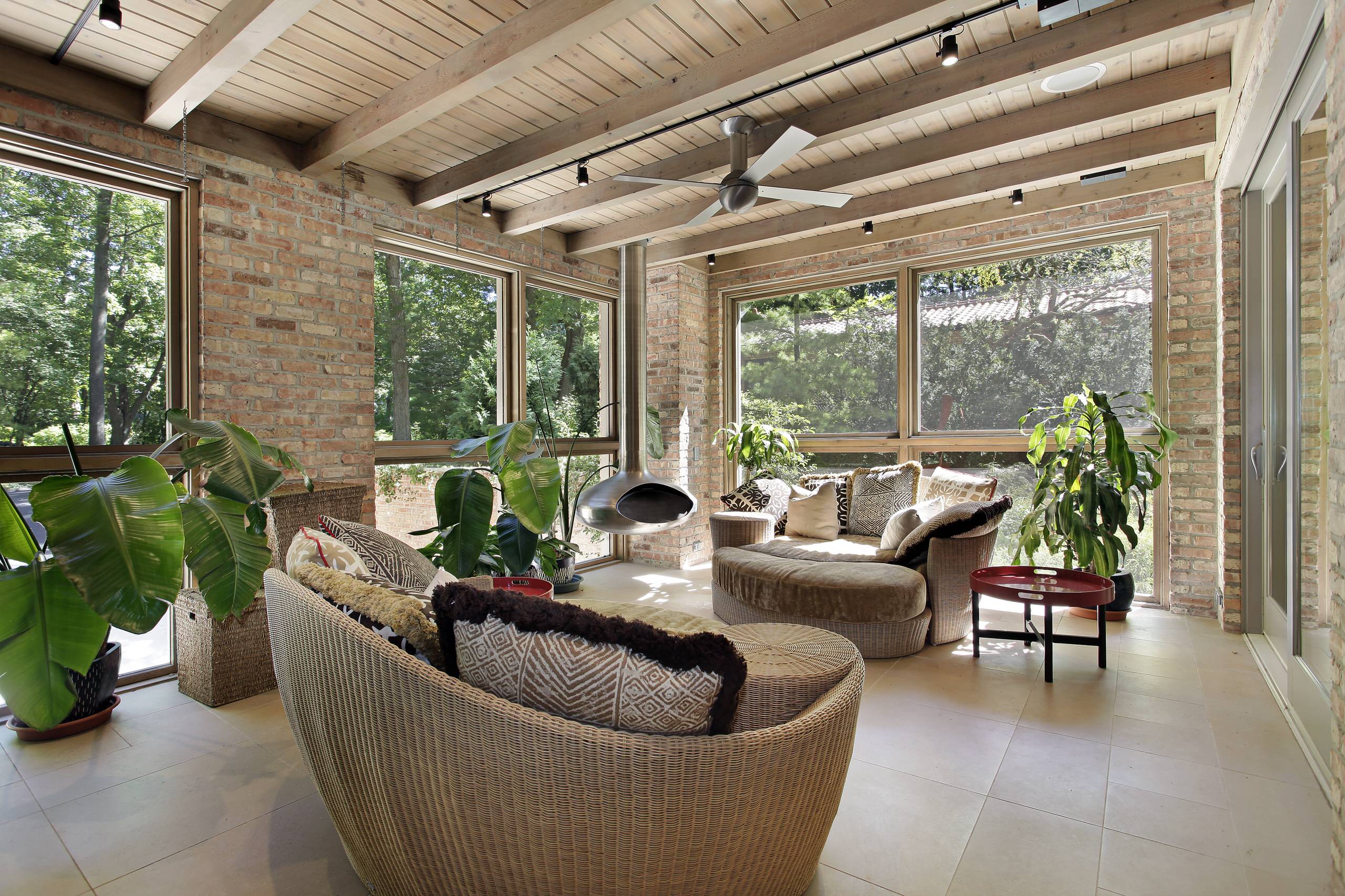 Sunroom with exposed brick walls, whitewashed ceiling beams, wicker loungers, and large tropical plants beside panoramic windows.