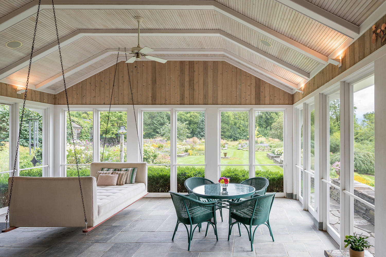 Bright sunroom featuring hanging daybed, teal wicker dining set, vaulted beadboard ceiling, and floor-to-ceiling windows overlooking garden.