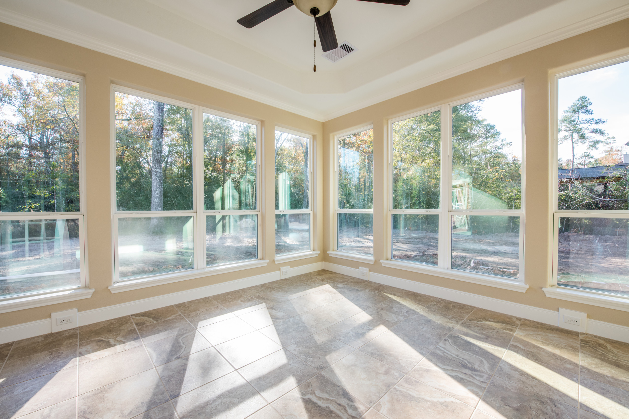 Bright sunroom with wraparound windows, marble-look tile flooring, and cream walls overlooking wooded landscape.