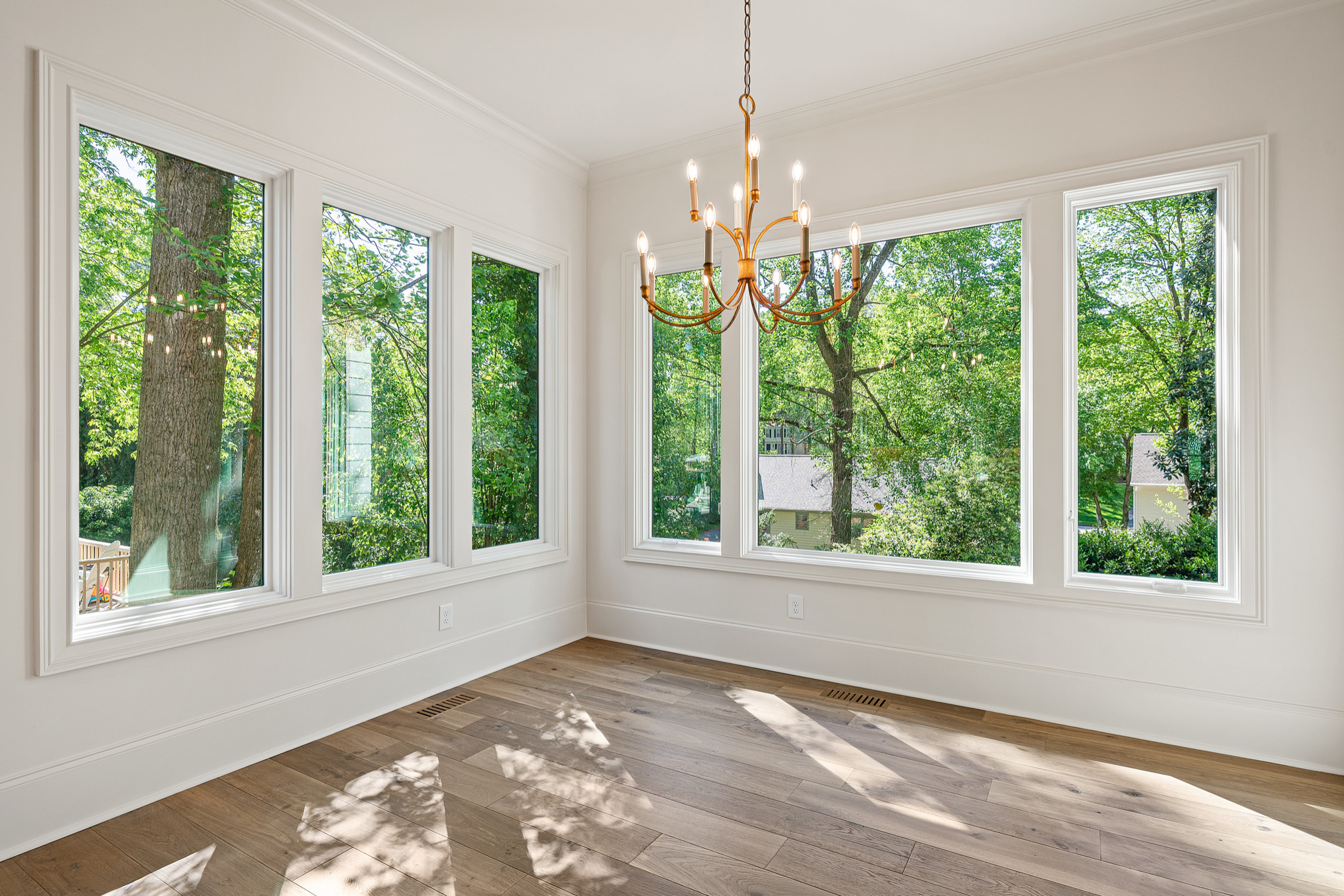 Bright corner room with panoramic windows, brass chandelier, white walls, and wide-plank oak flooring showcasing forest views.