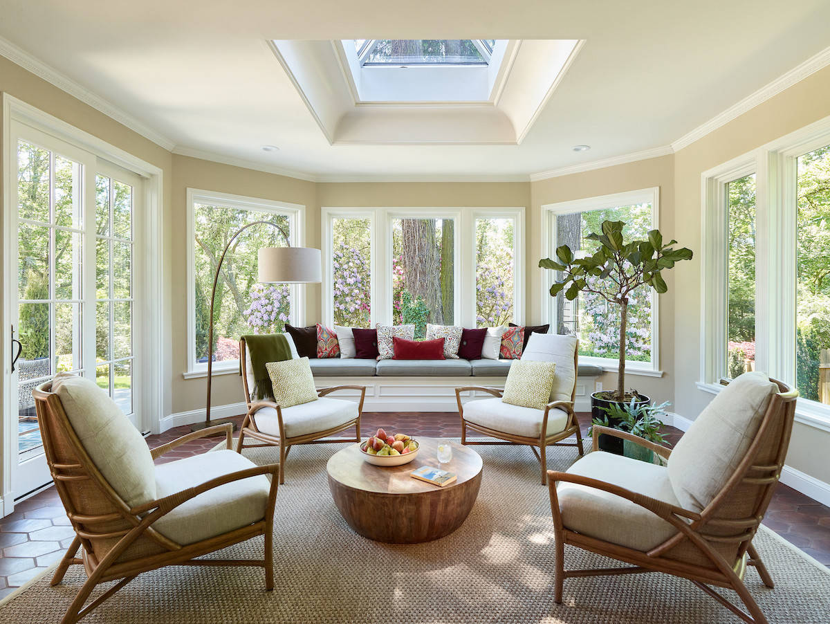 Bright sunroom with wraparound windows, built-in bench, midcentury chairs, round wooden coffee table, and skylight above.