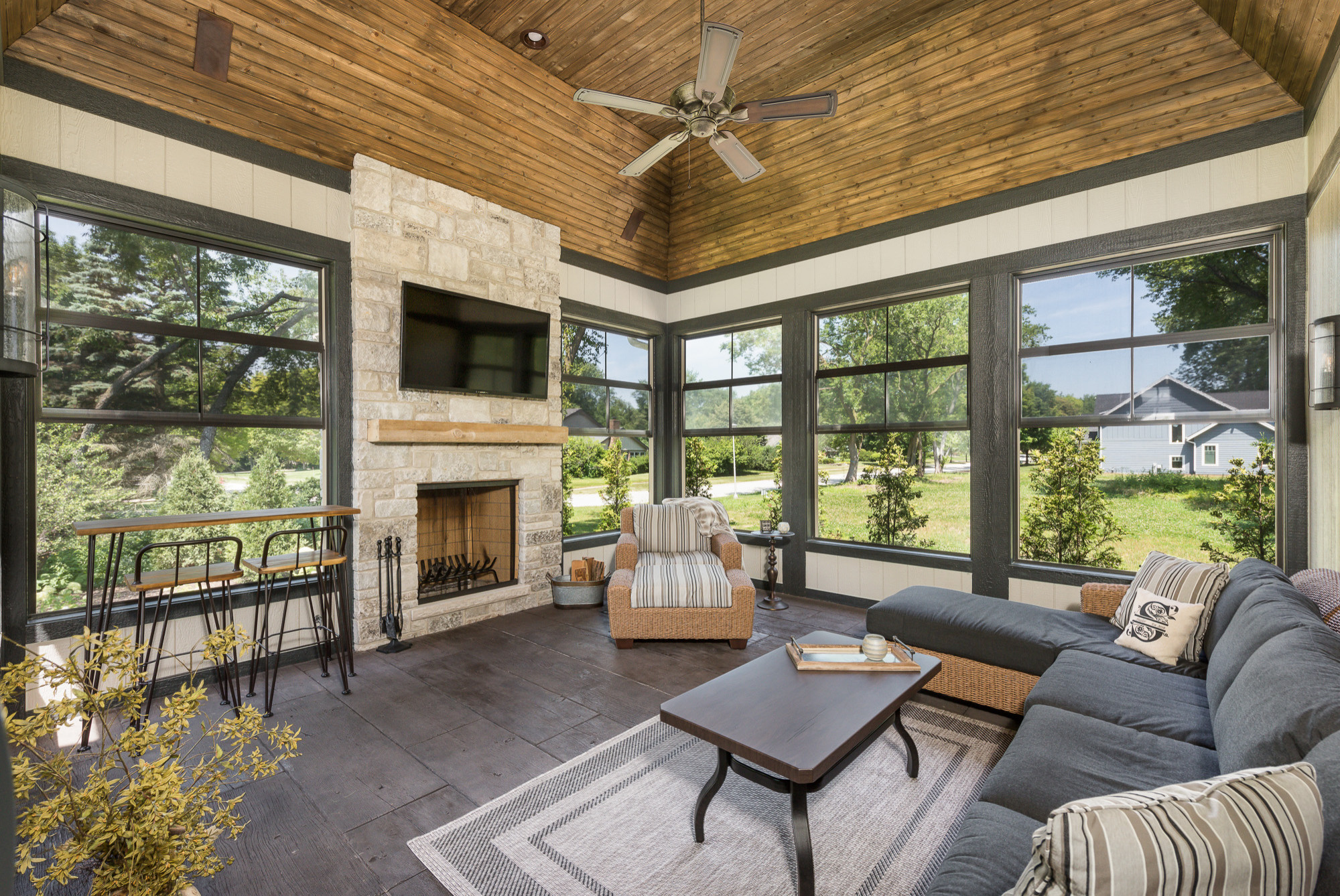 Contemporary sunroom featuring stone fireplace, wooden ceiling, panoramic windows, sectional sofa, and bar-height seating area.