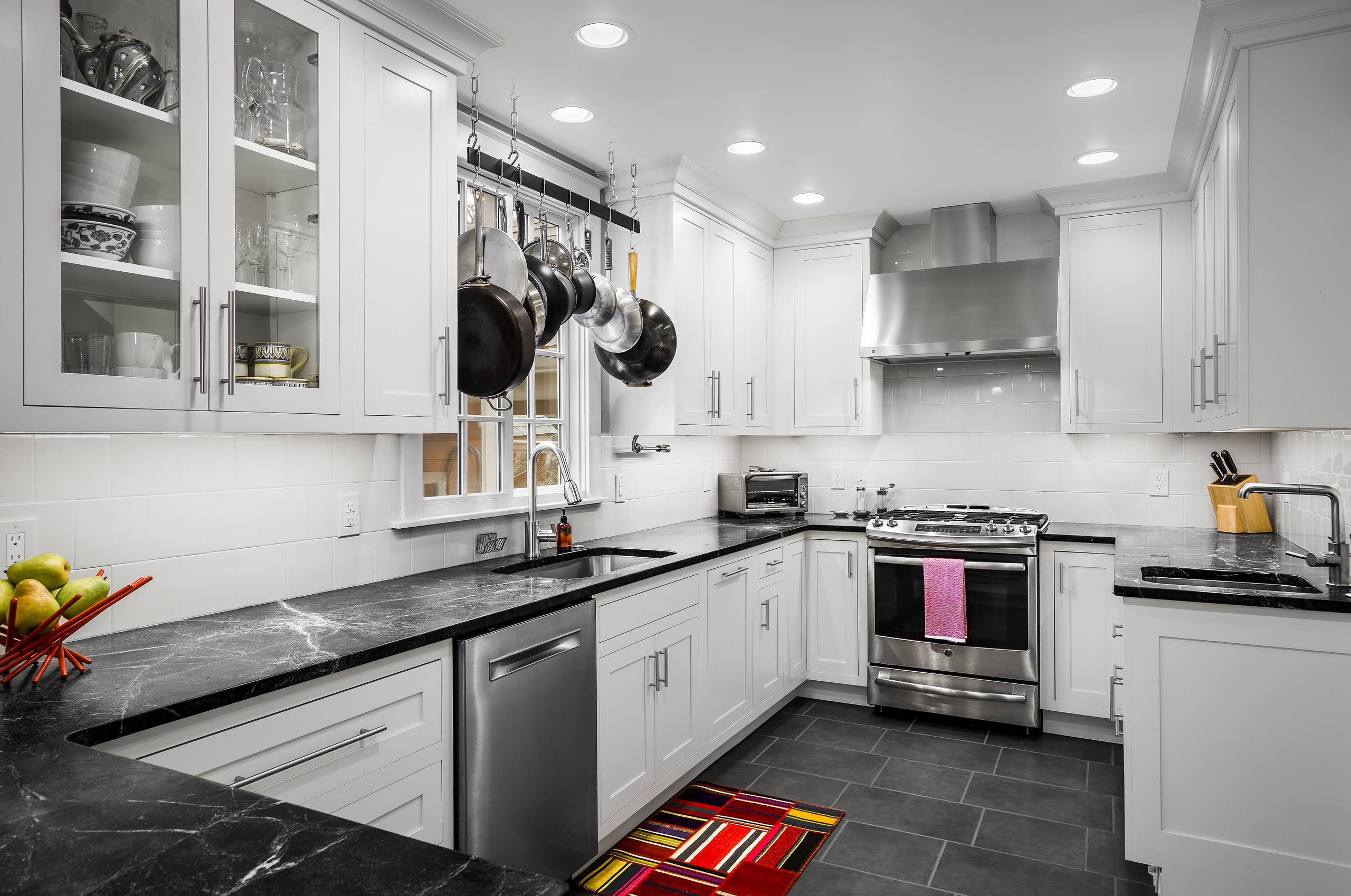 L-shaped white kitchen with black marble countertops, hanging cookware, stainless appliances, and gray tile flooring.