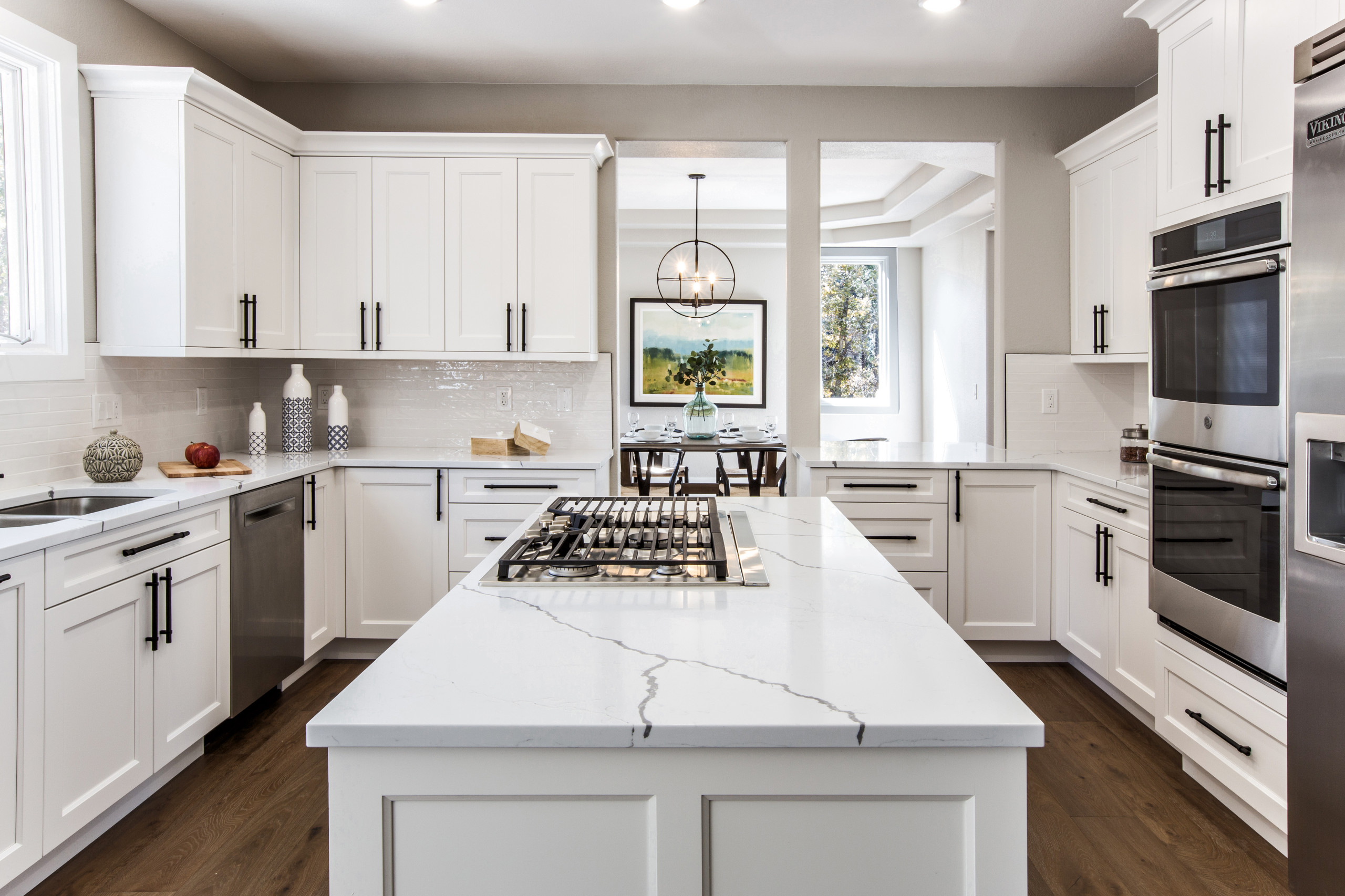 Modern white kitchen featuring marble island with gas cooktop, black hardware, stainless appliances, and hardwood flooring.