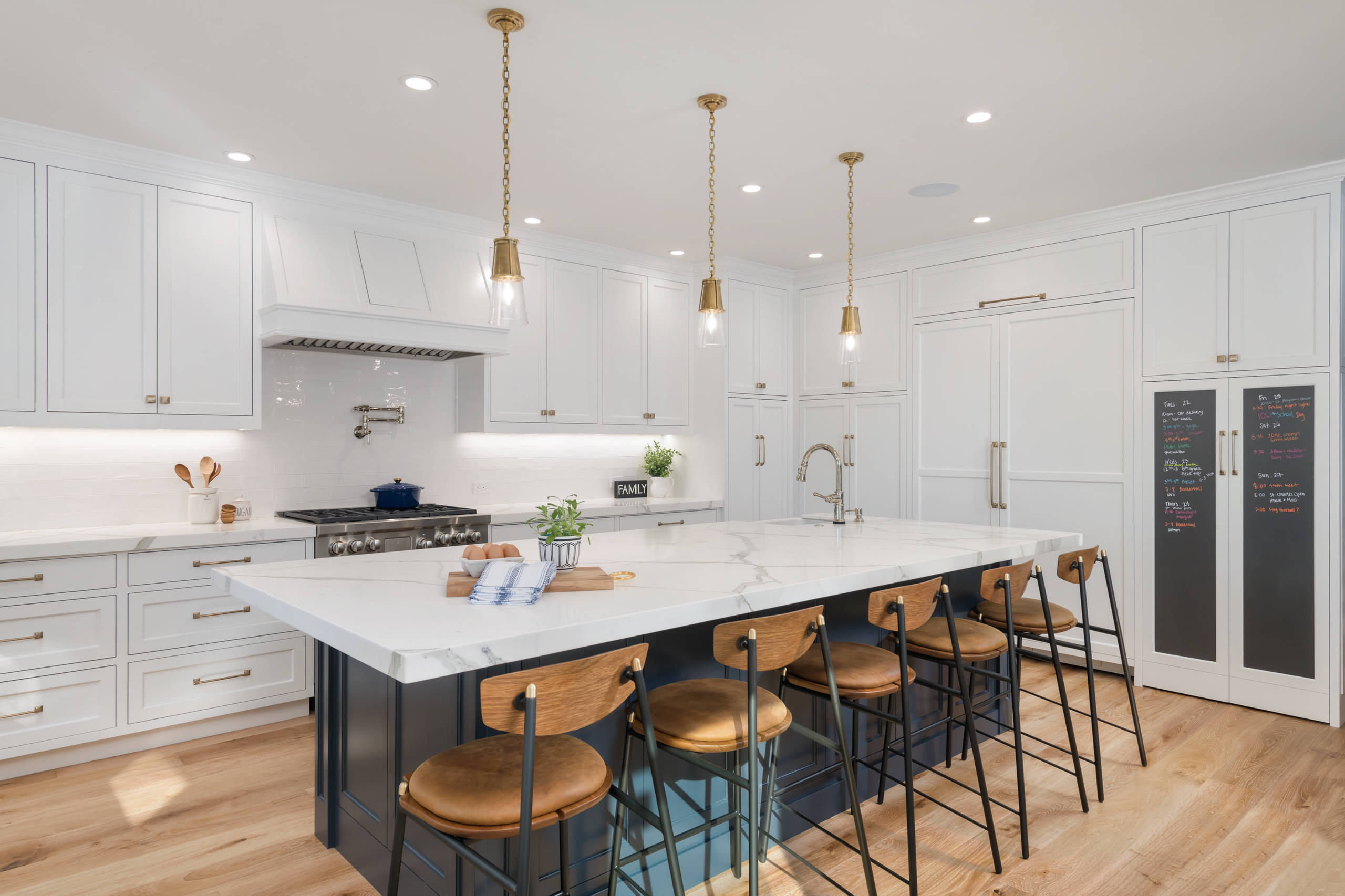 Bright white kitchen with marble waterfall island, brass pendant lights, leather barstools, and navy blue accents.