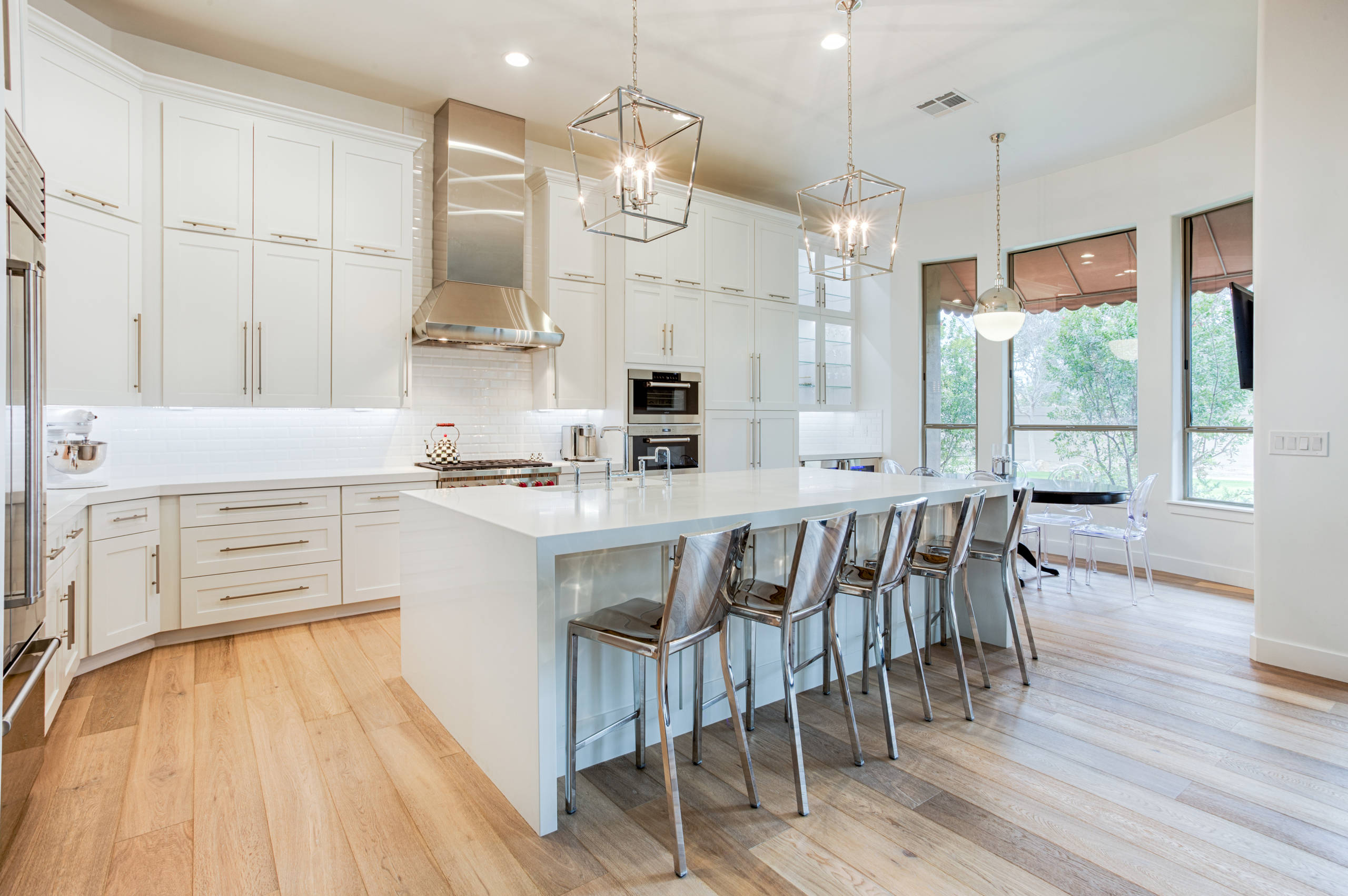 Bright white kitchen featuring floor-to-ceiling cabinets, stainless steel appliances, geometric pendant lights, and chrome barstools along white island.