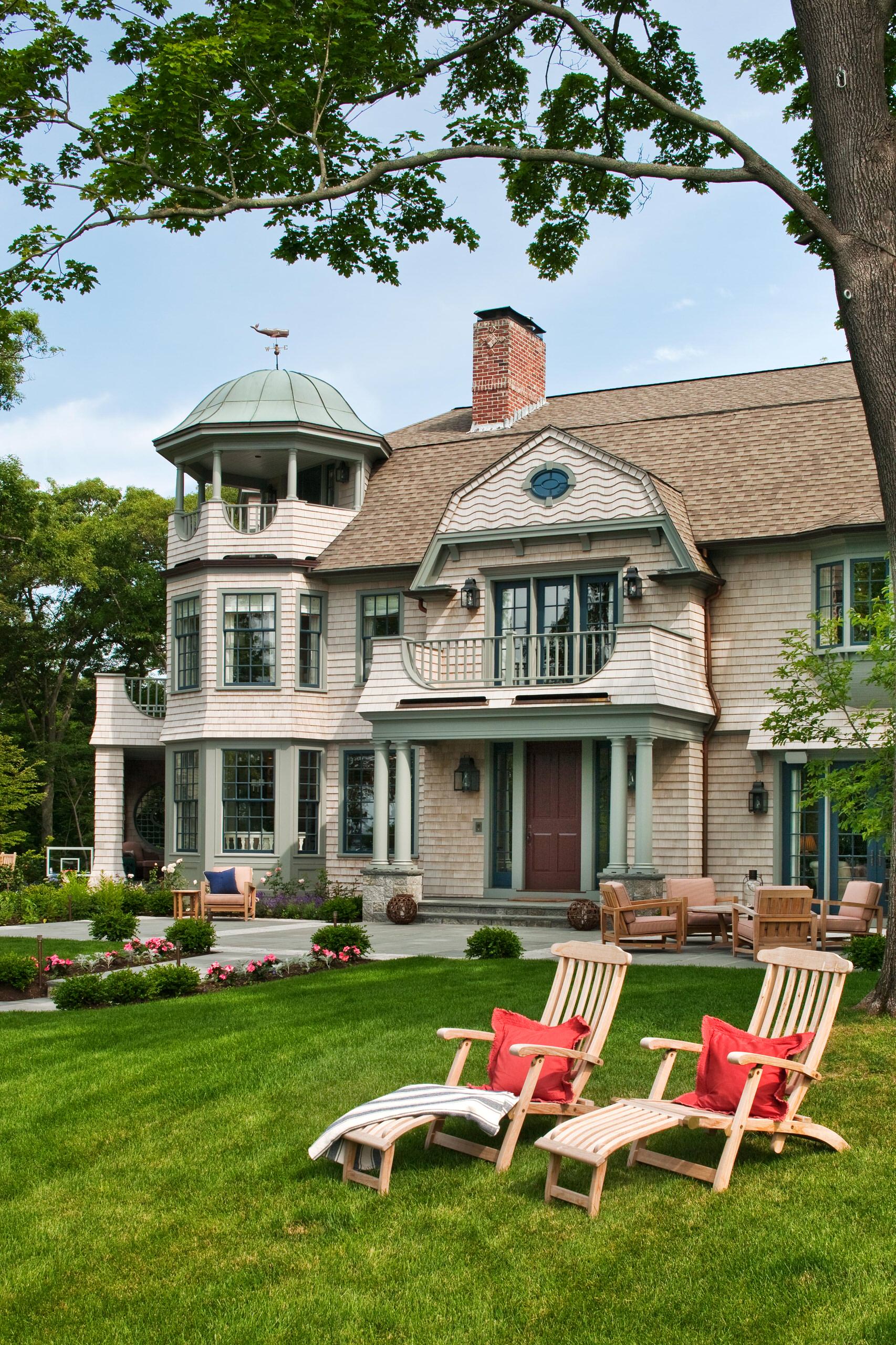 Shingle-style mansion featuring copper-domed tower, wraparound porch, and manicured lawn with teak loungers and garden beds.