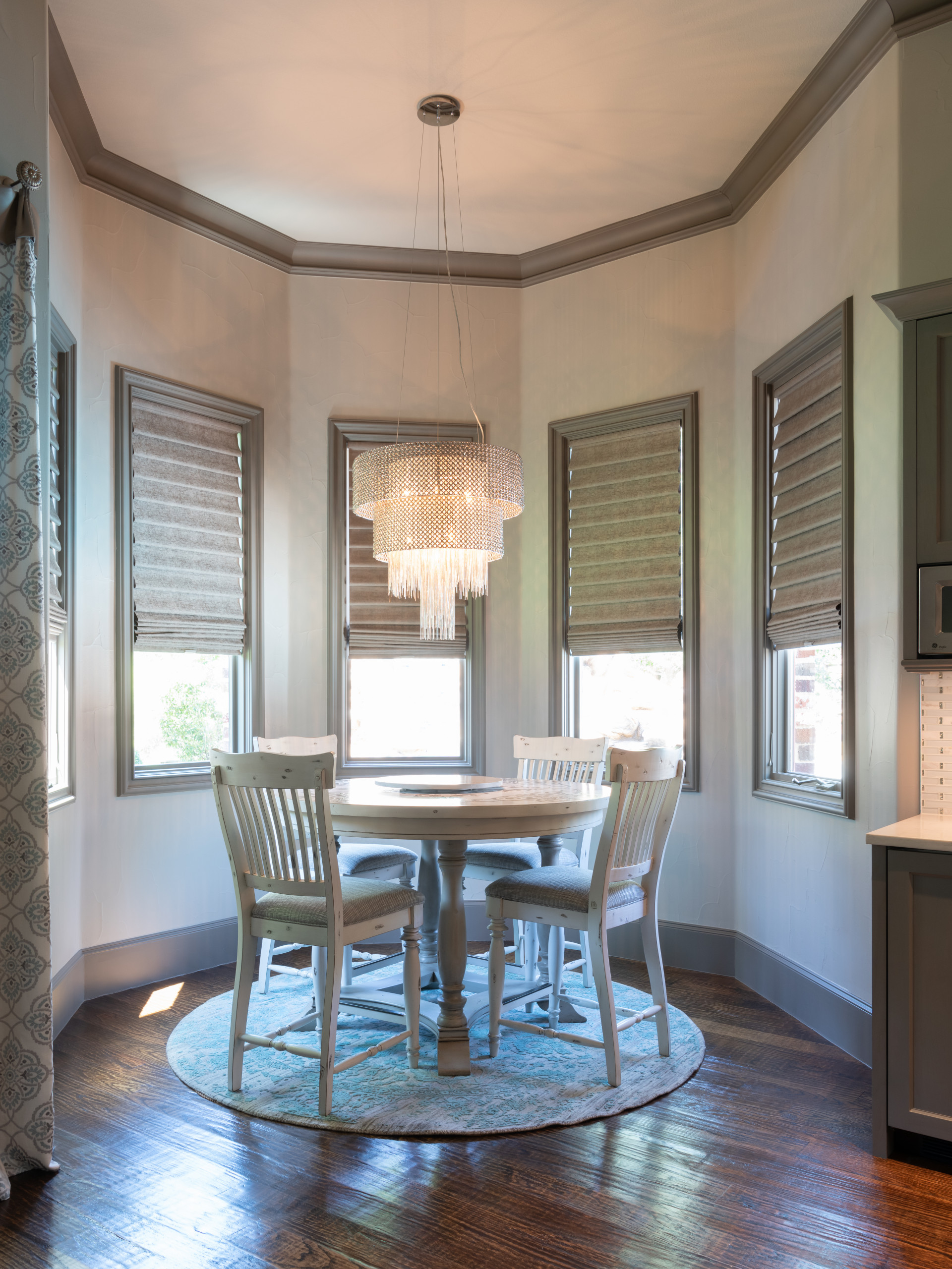 Octagonal breakfast nook with tiered crystal chandelier, round white table, four windows with grey Roman shades, and hardwood floors.
