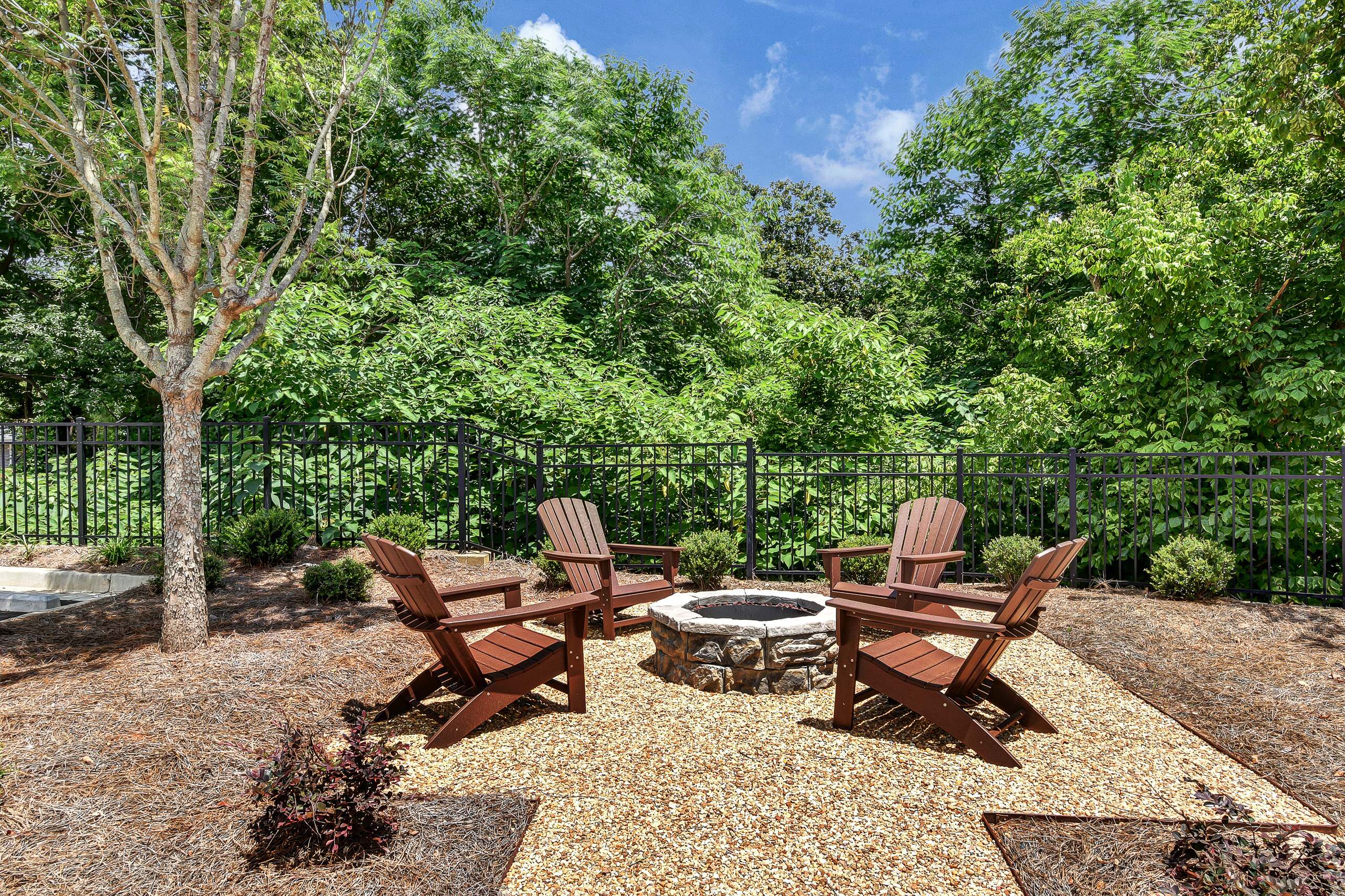 Circular stone fire pit surrounded by four brown Adirondack chairs on gravel patio, bordered by black metal fence and lush greenery.